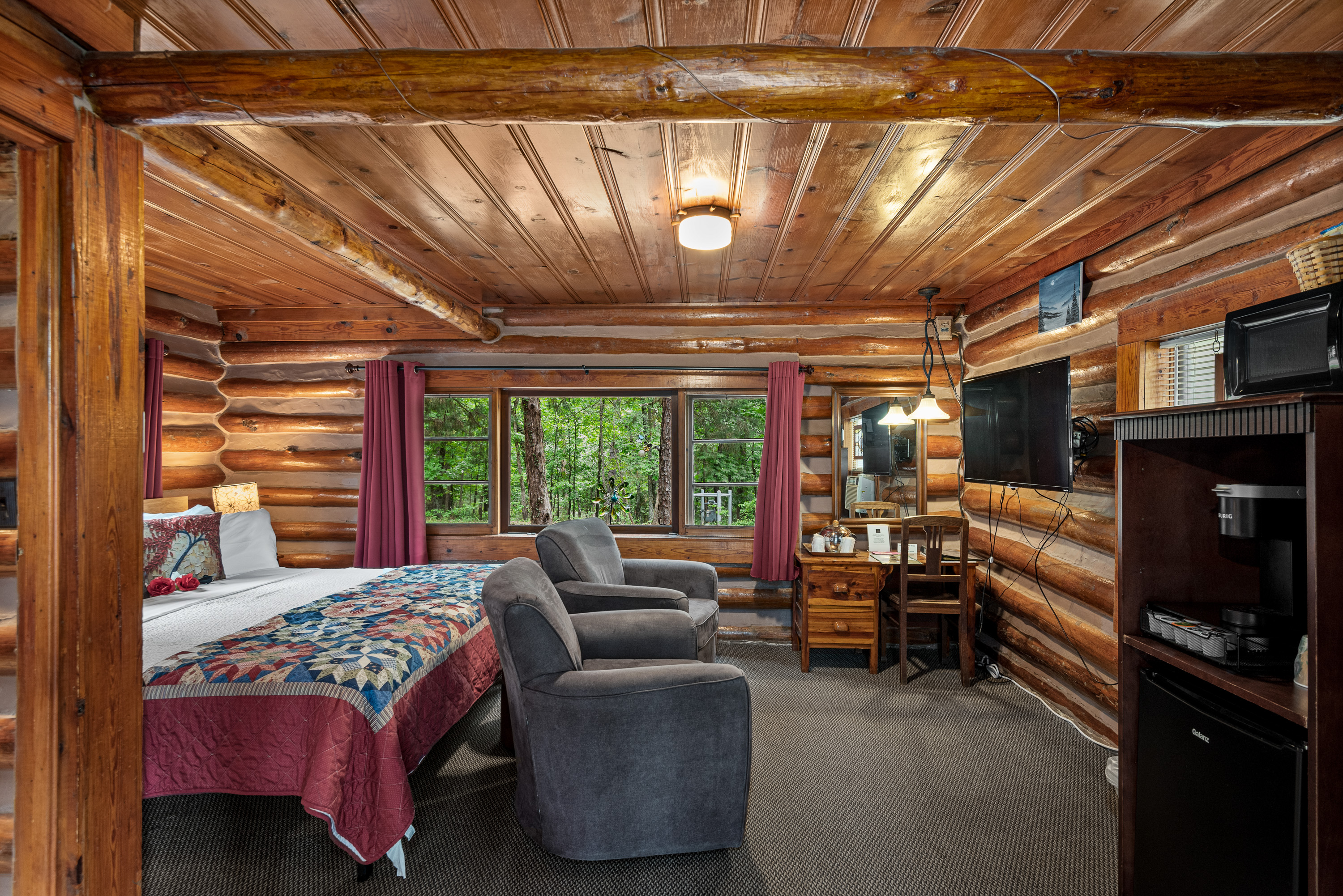 A rustic log cabin bedroom with a king-size bed, two armchairs, a small desk, a television, and a small refrigerator and microwave.