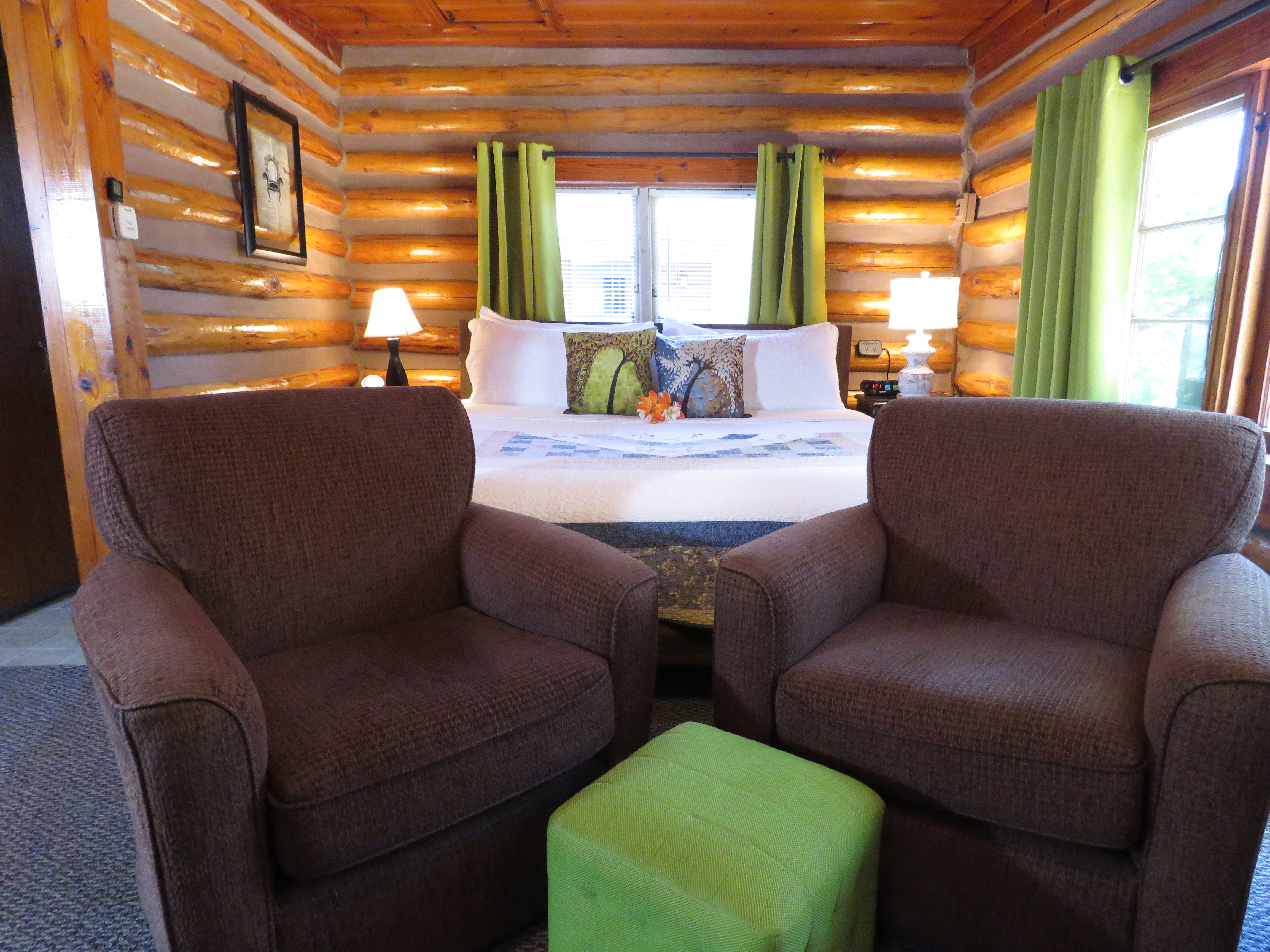 A king-size bed, a pair of armchairs, and a green ottoman are in a cozy log cabin bedroom. The rustic walls and ceiling are made of natural wood.