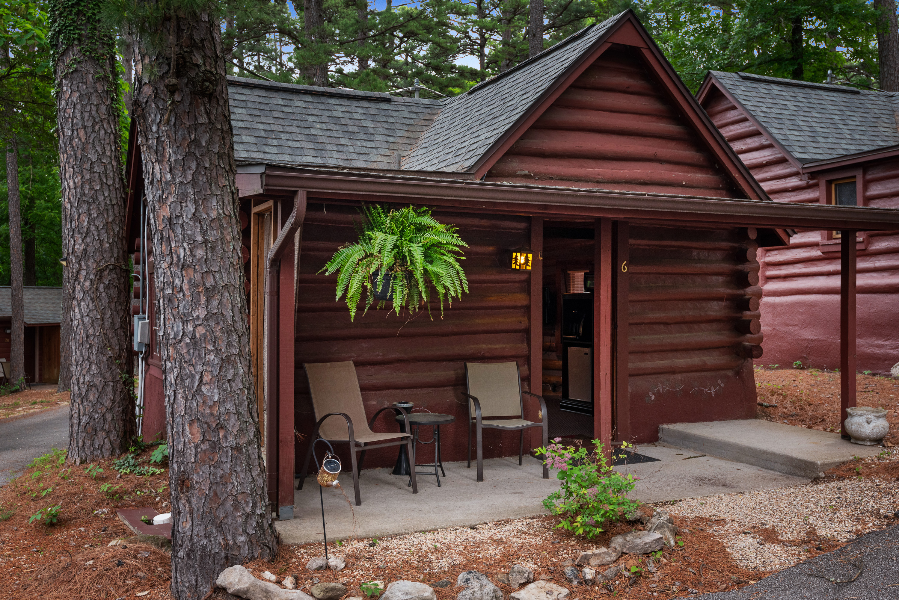 A charming log cabin is nestled among trees, with a covered porch holding two chairs and a small table. A fern in a hanging basket hangs on the porch.