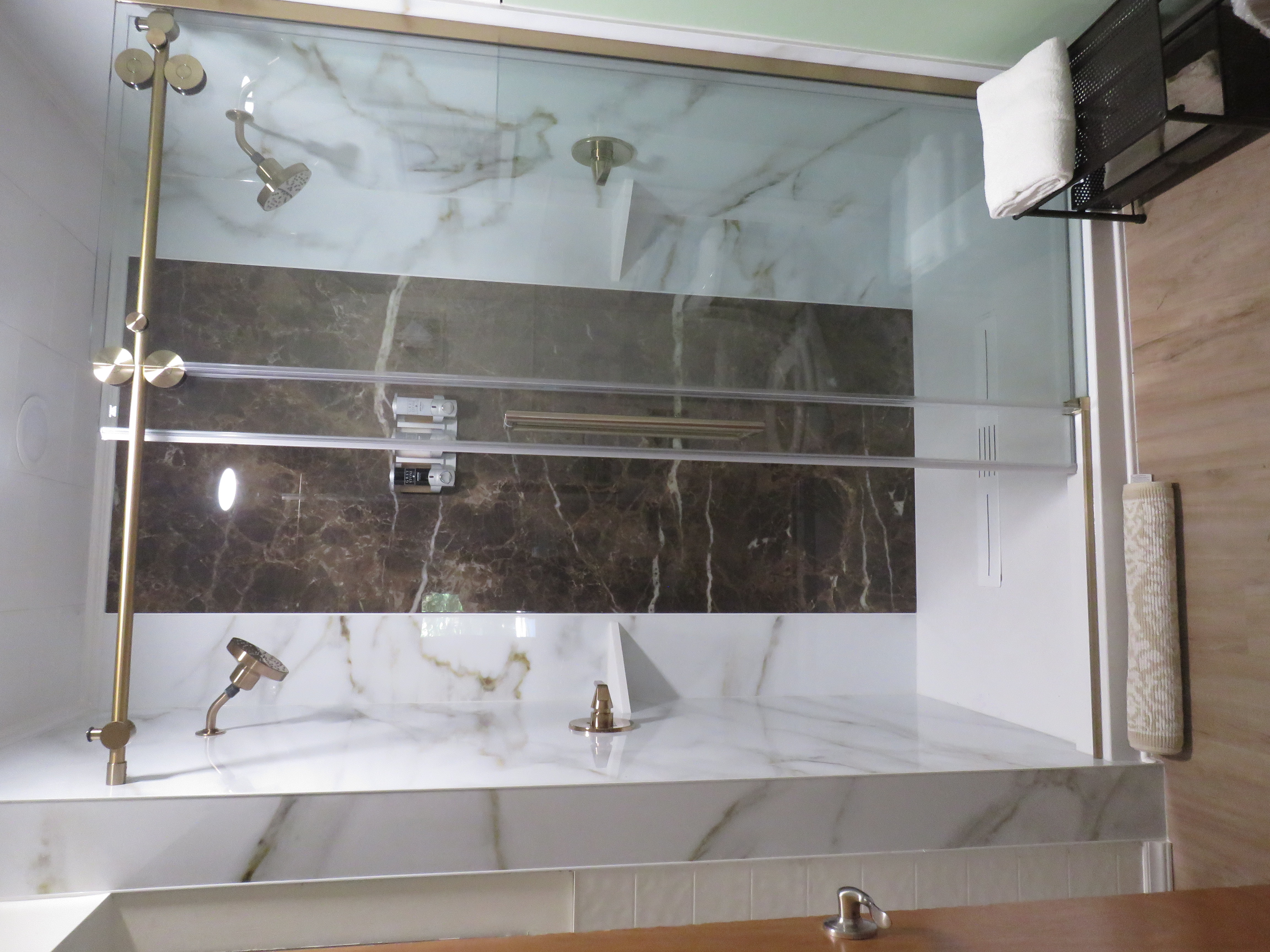 Modern shower with glass doors, dual shower heads, marble walls, and a dark brown accent panel. A small bench with folded towels and a rolled mat are in front of the entrance.