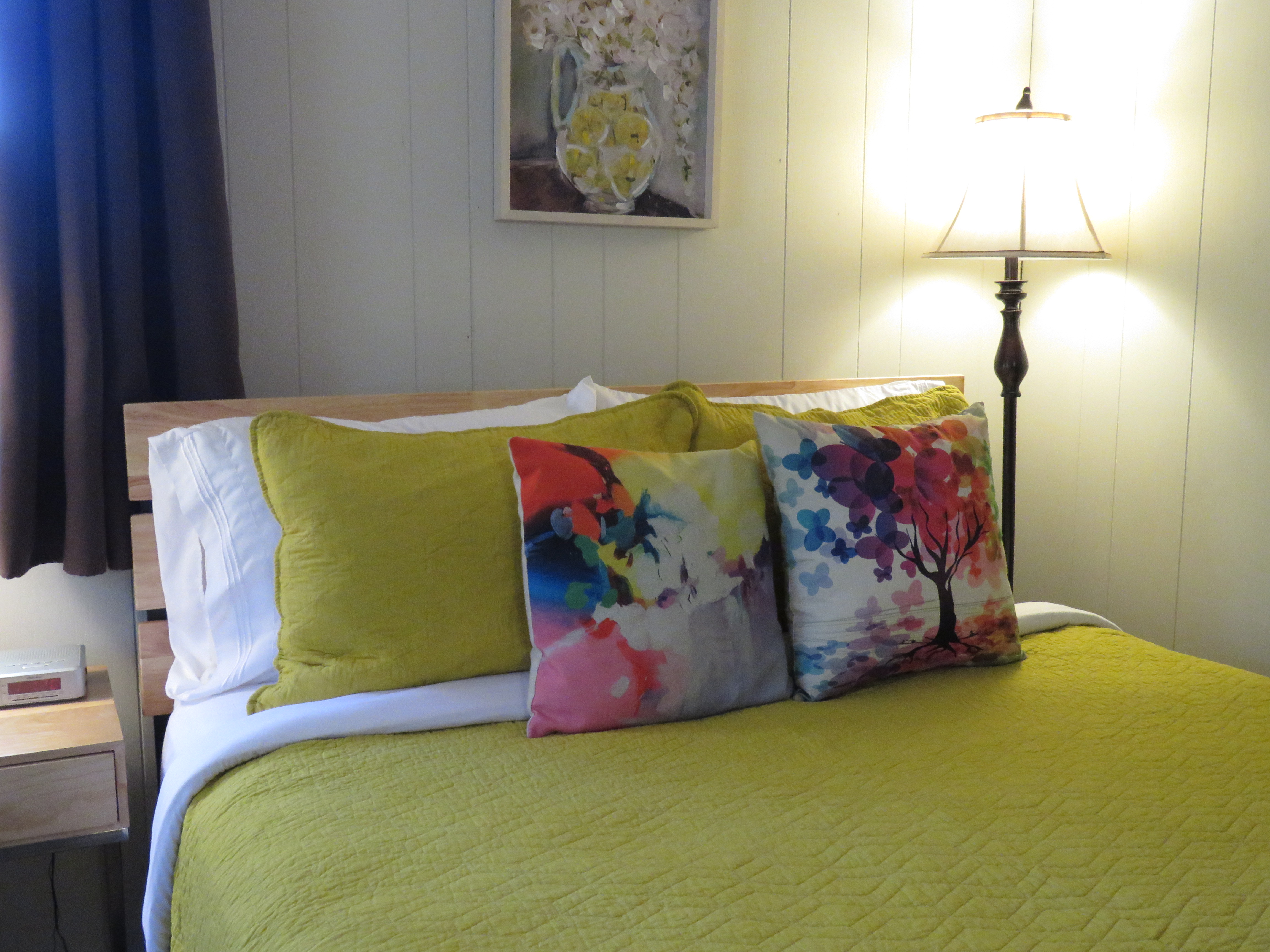 A queen bed with a bright yellow comforter and three pillows. The pillows include a solid yellow one and two with colorful, modern patterns.