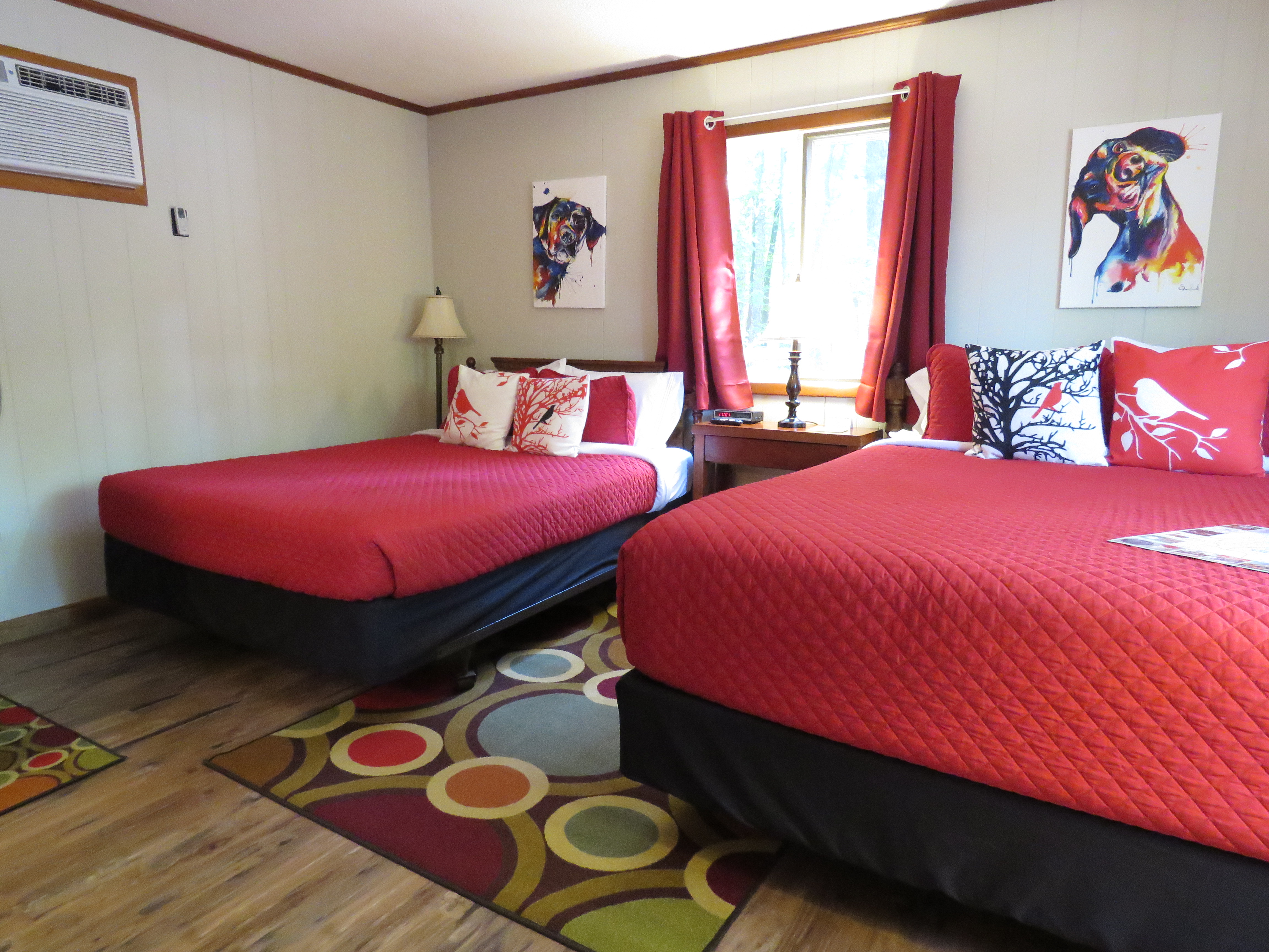 A cozy room features two beds with bright red quilts and dog-themed art on the walls. A colorful, patterned rug is on the floor.