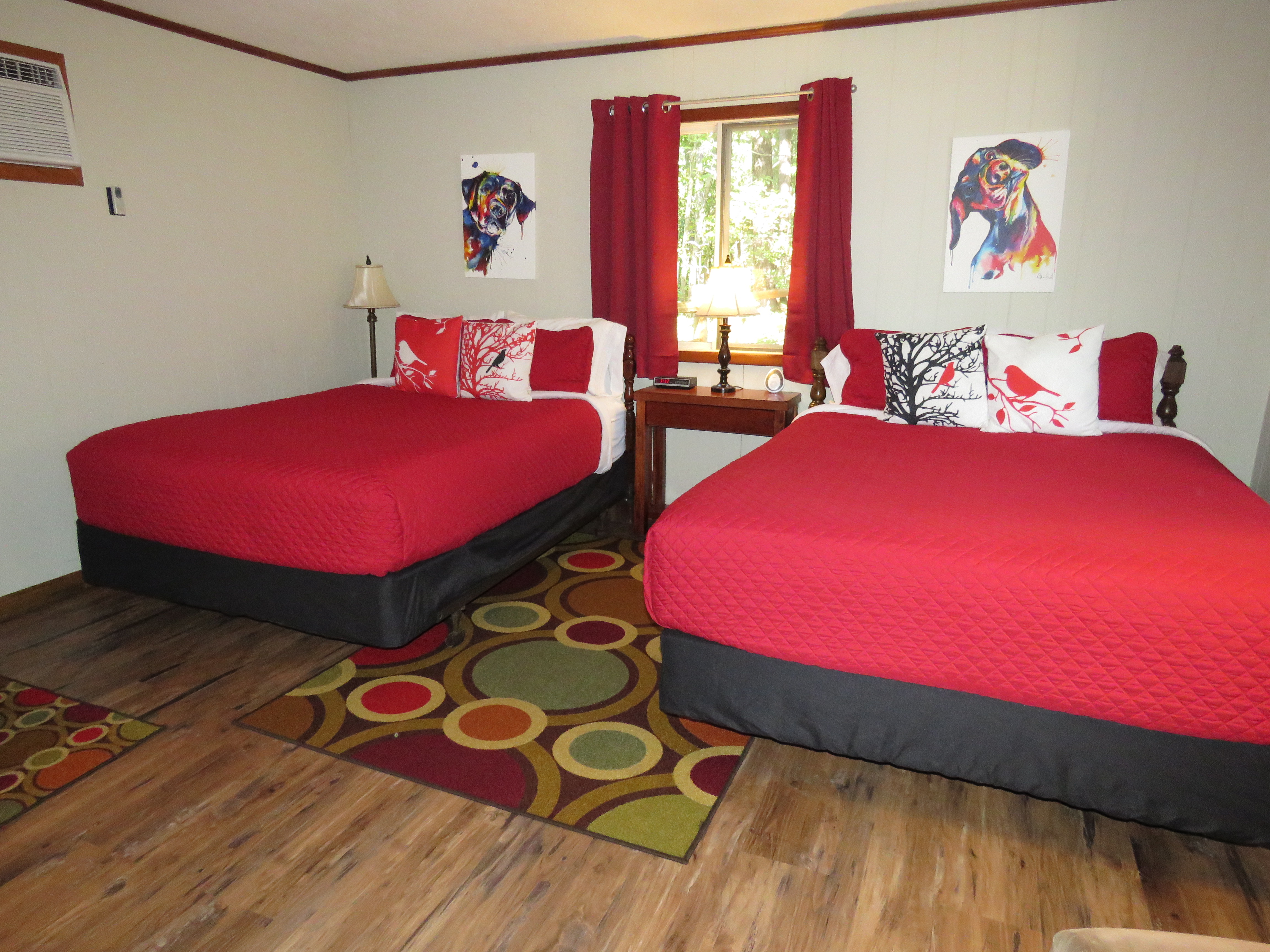 A cozy room features two beds with bright red quilts and dog-themed art on the walls. A colorful, patterned rug is on the floor.