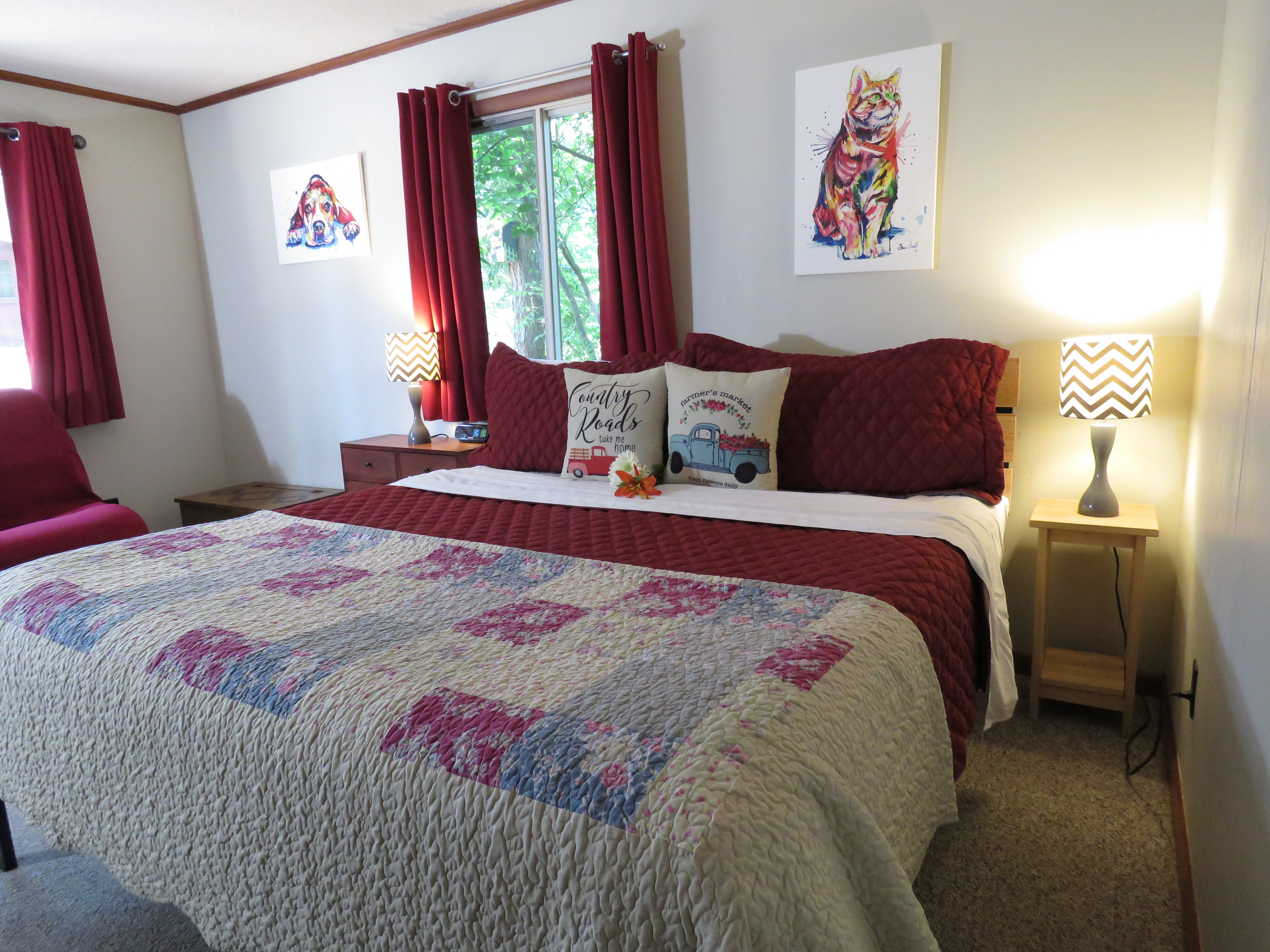 A bedroom features a king-size bed with a red comforter and a patchwork quilt. The walls have a neutral color, with pet-themed art and red curtains on the windows.