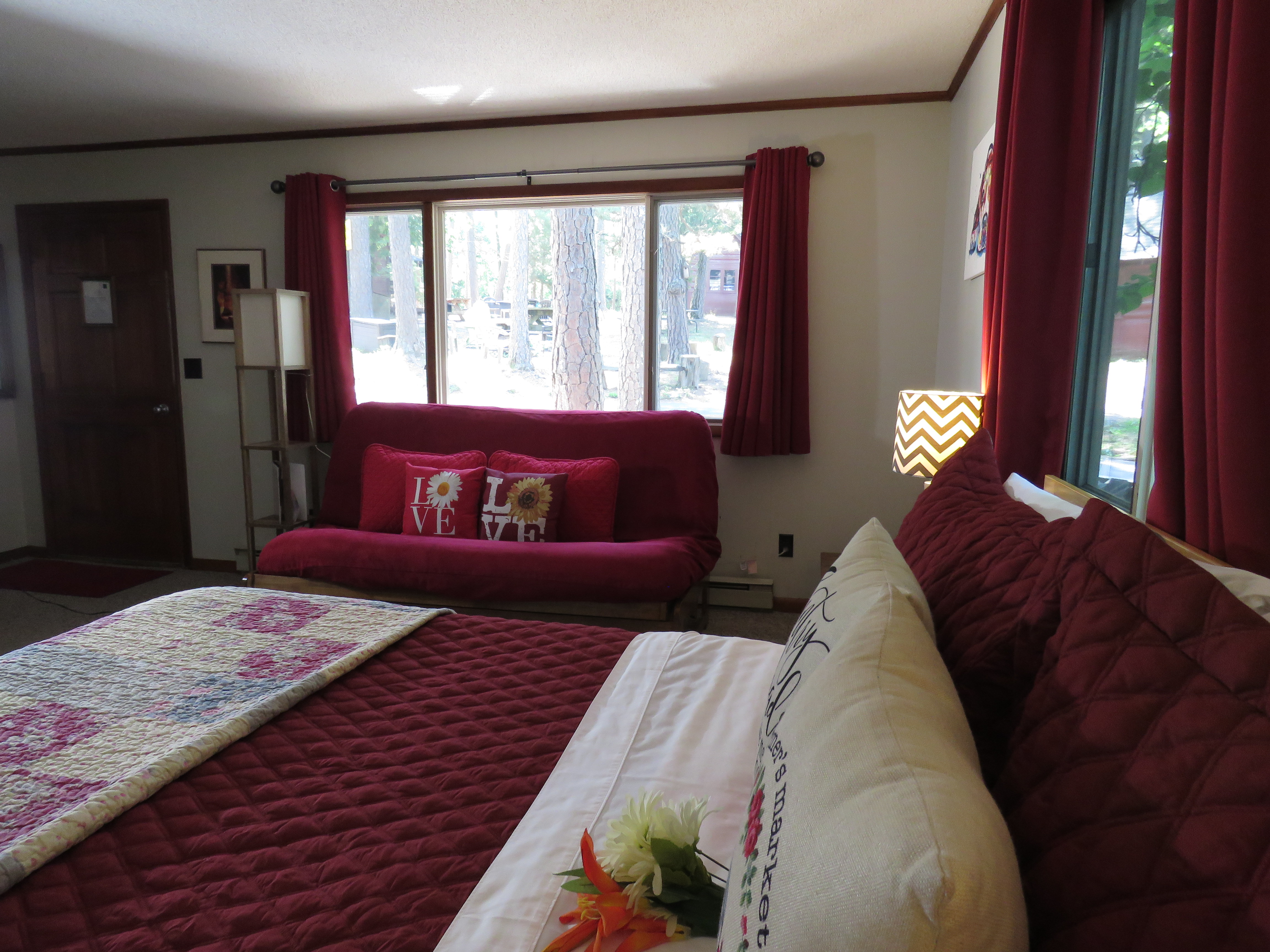 A bedroom with a dark red comforter on the king bed, red curtains on the windows, and a red futon