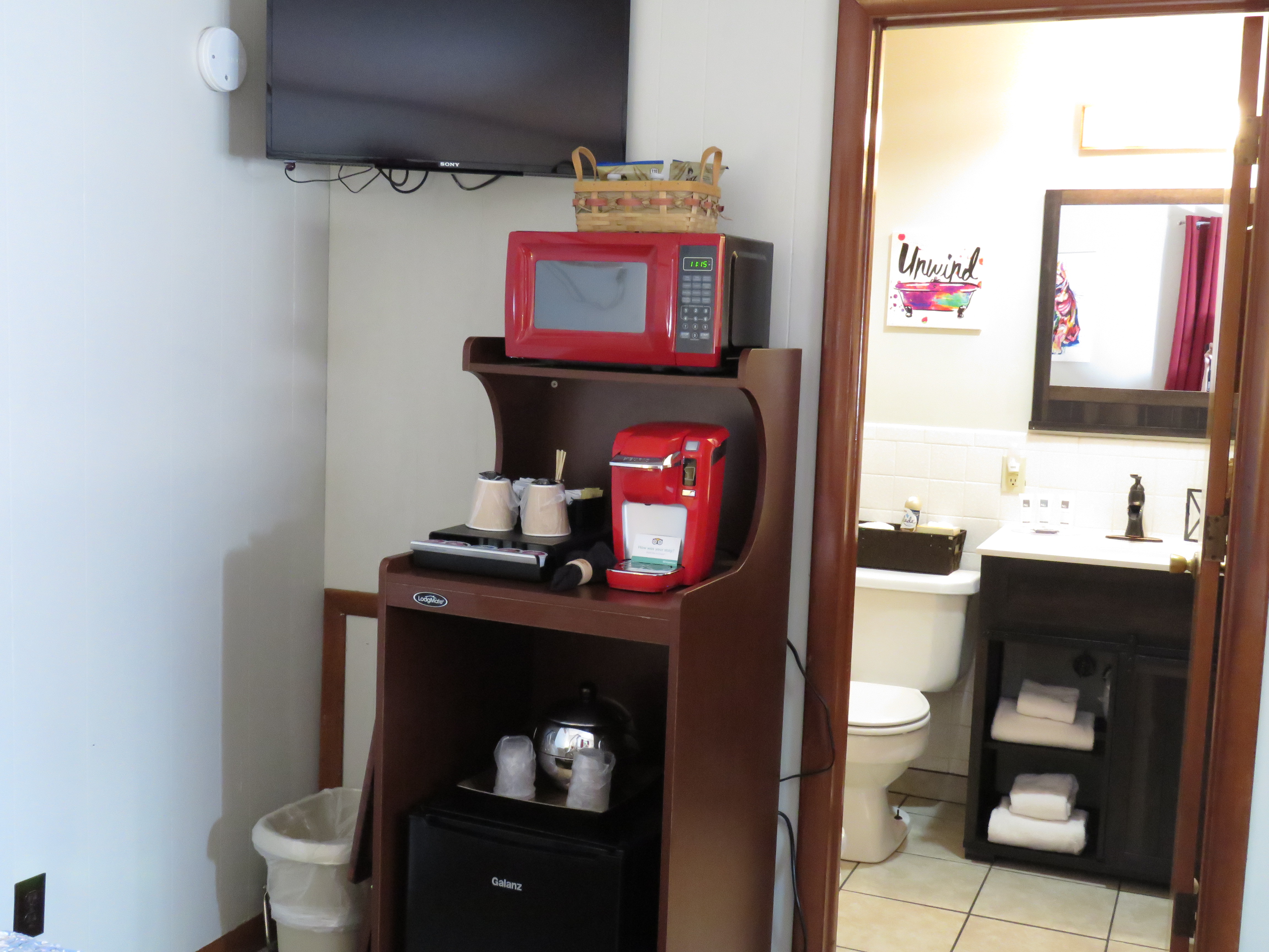 A beverage station with a red microwave, a red single-serve coffee maker, and a mini-fridge are on a dark wood stand in a bedroom. An open doorway leads to a small bathroom.