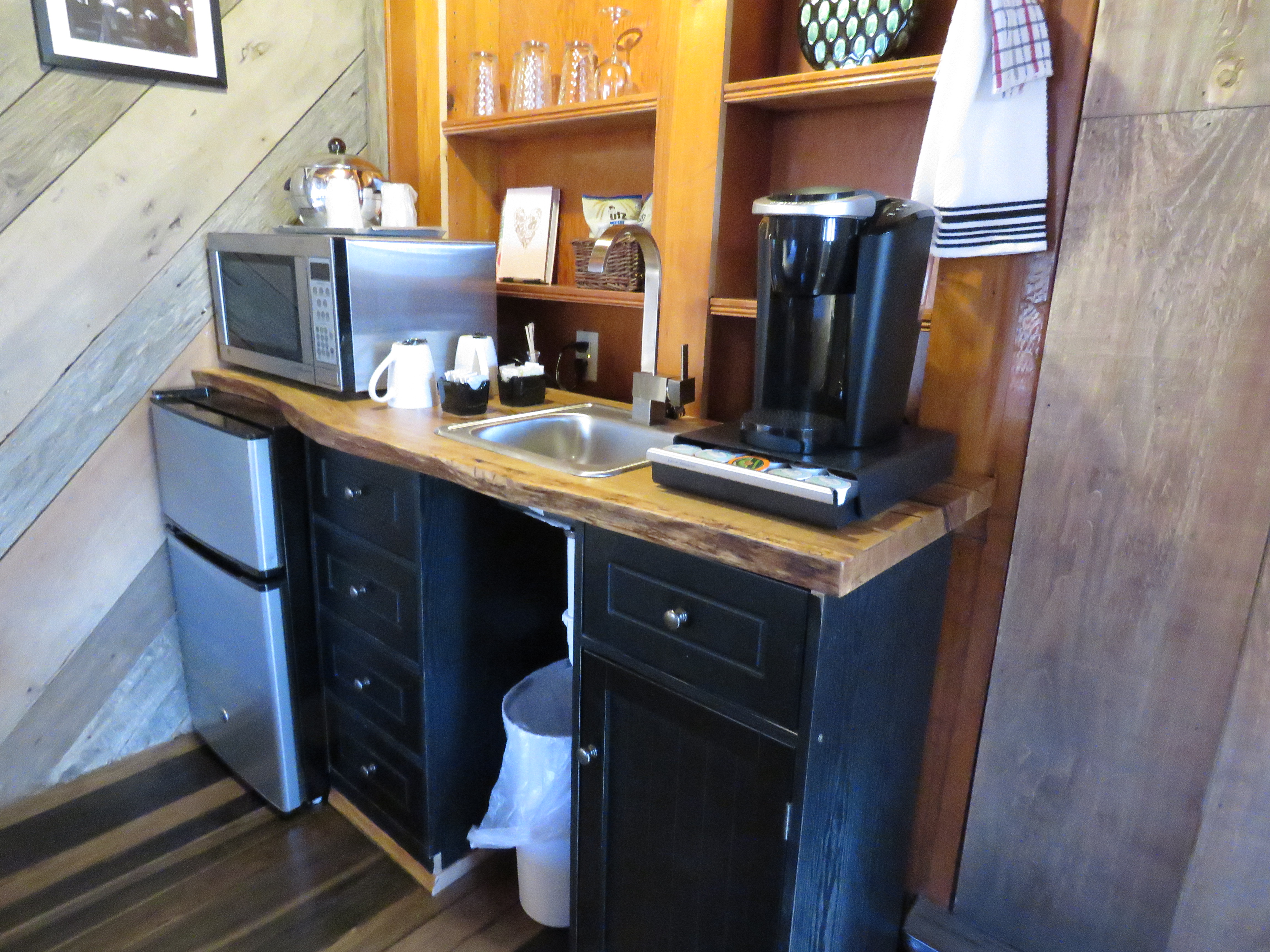 A small, rustic kitchenette with a live-edge countertop and black cabinets. The space has a microwave, a mini-fridge, and a coffee maker.