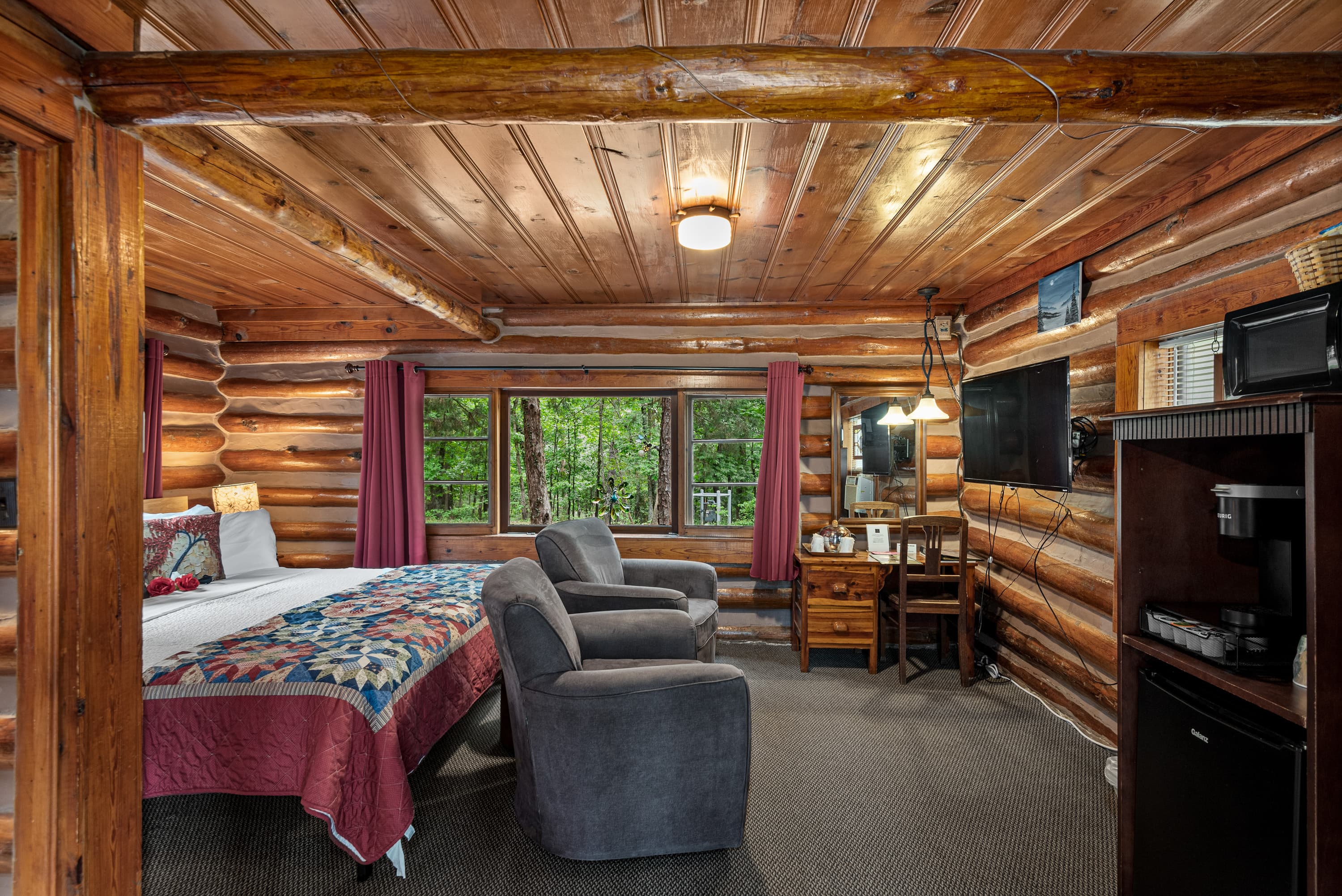A rustic log cabin bedroom with a king-size bed, two armchairs, a small desk, a television, and a small refrigerator and microwave.