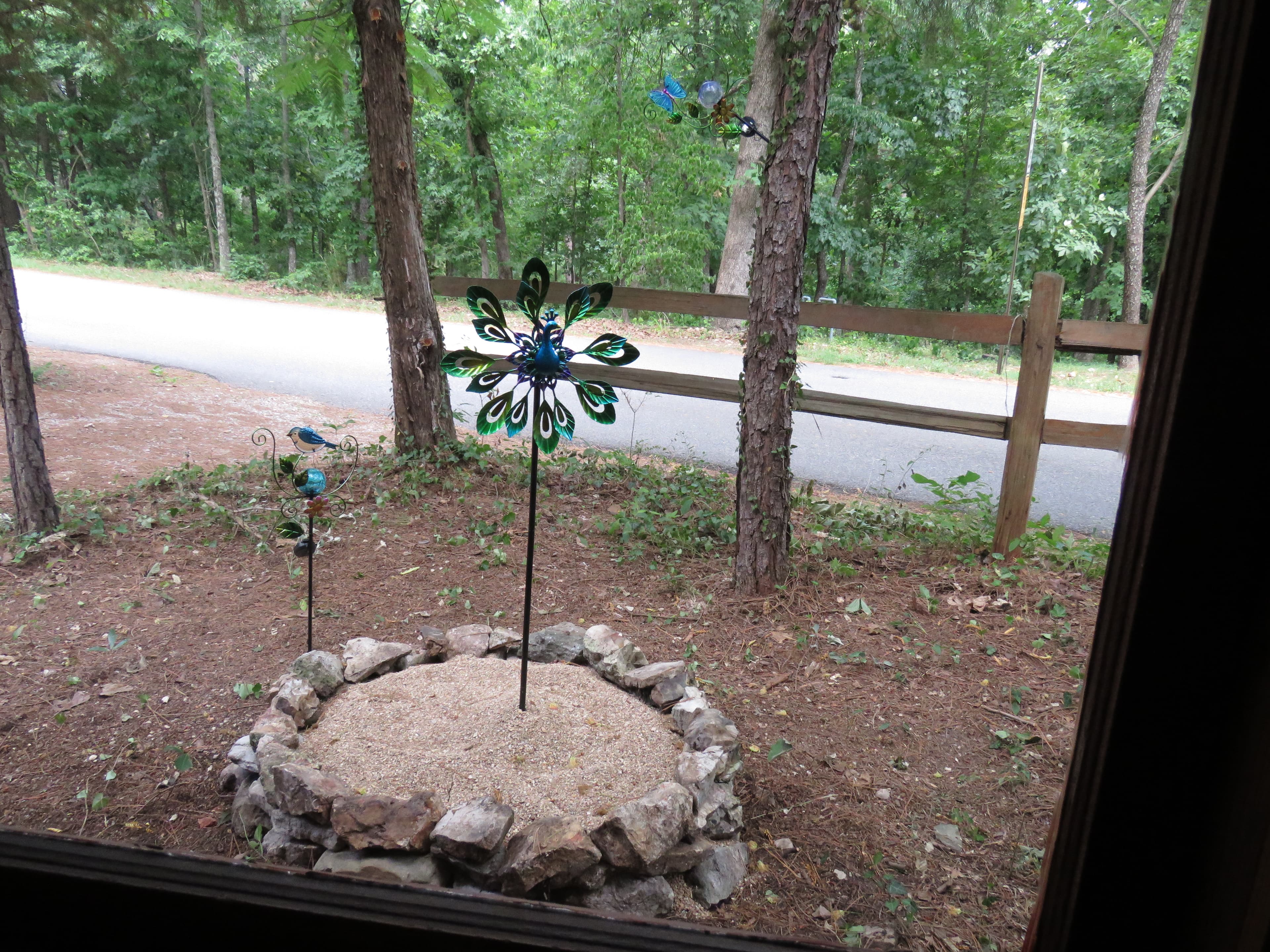 A garden ornament of a blue peacock pinwheel sits in a rock-lined bed of light-colored gravel. A wooden fence and trees are in the background.