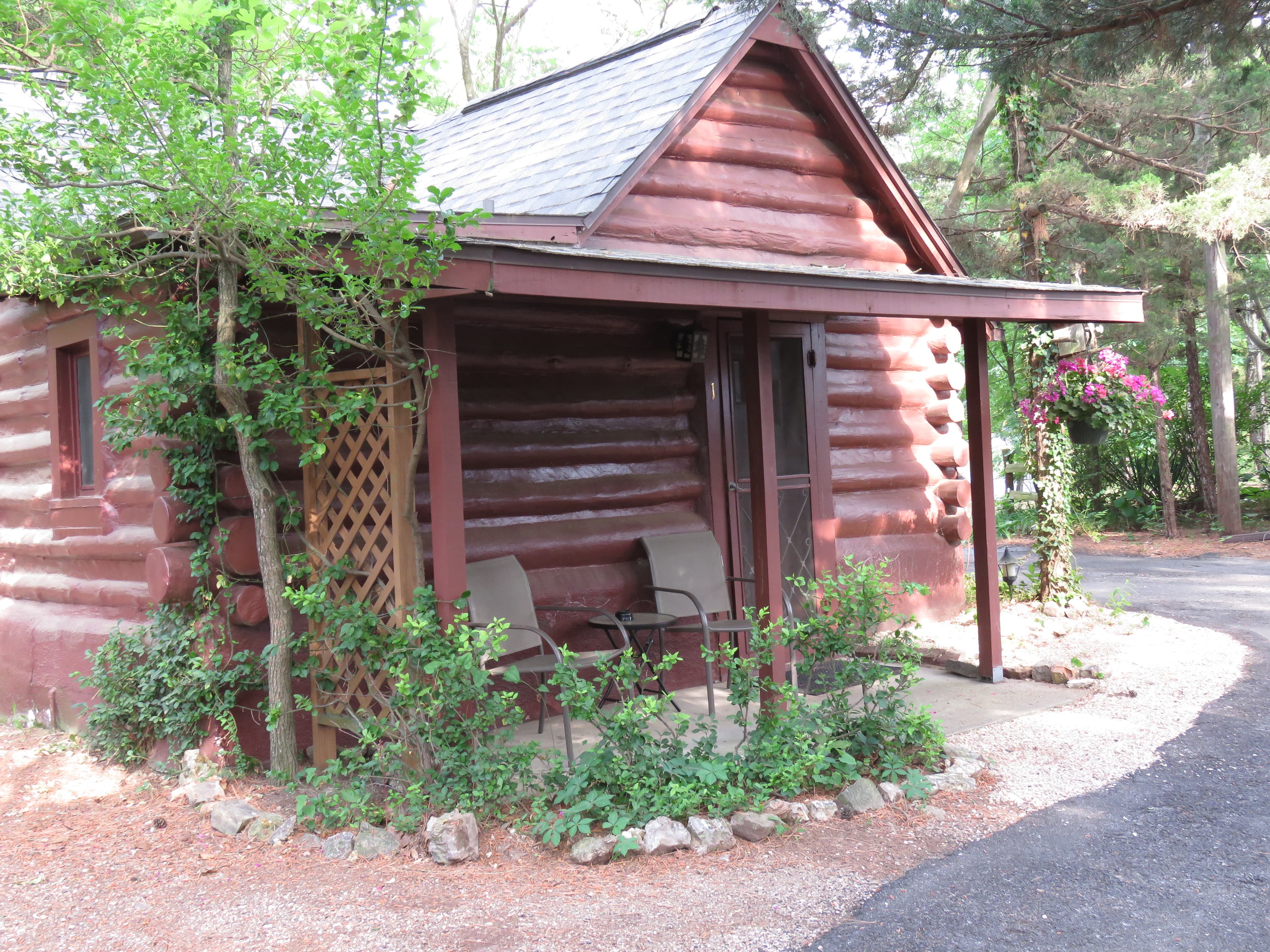A cozy, small log cabin with a covered porch holding two chairs. The cabin is surrounded by a small garden with a trellis and lush greenery.