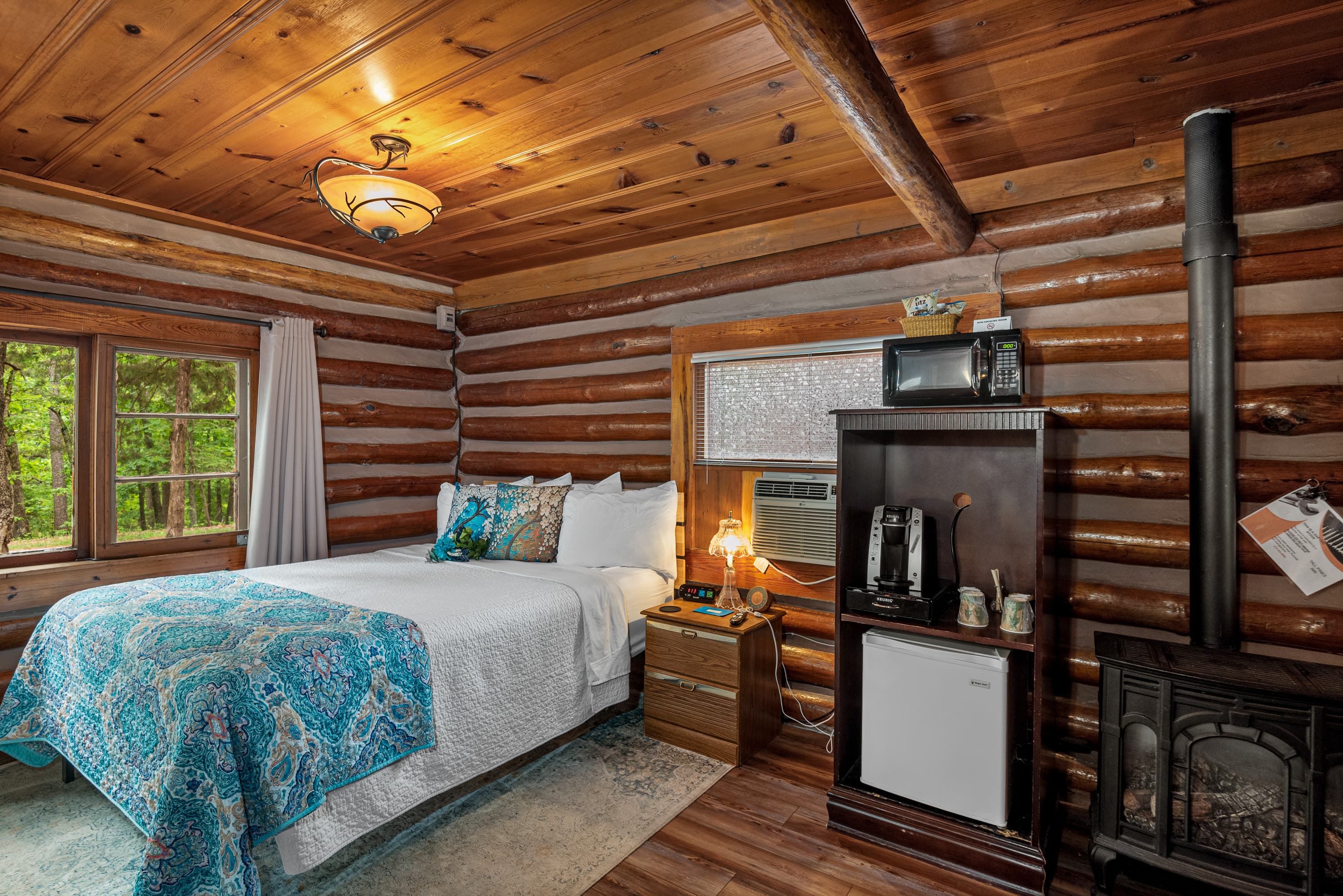 A rustic log cabin bedroom with a queen bed, a wood stove, and a small beverage center with a microwave and mini-fridge.