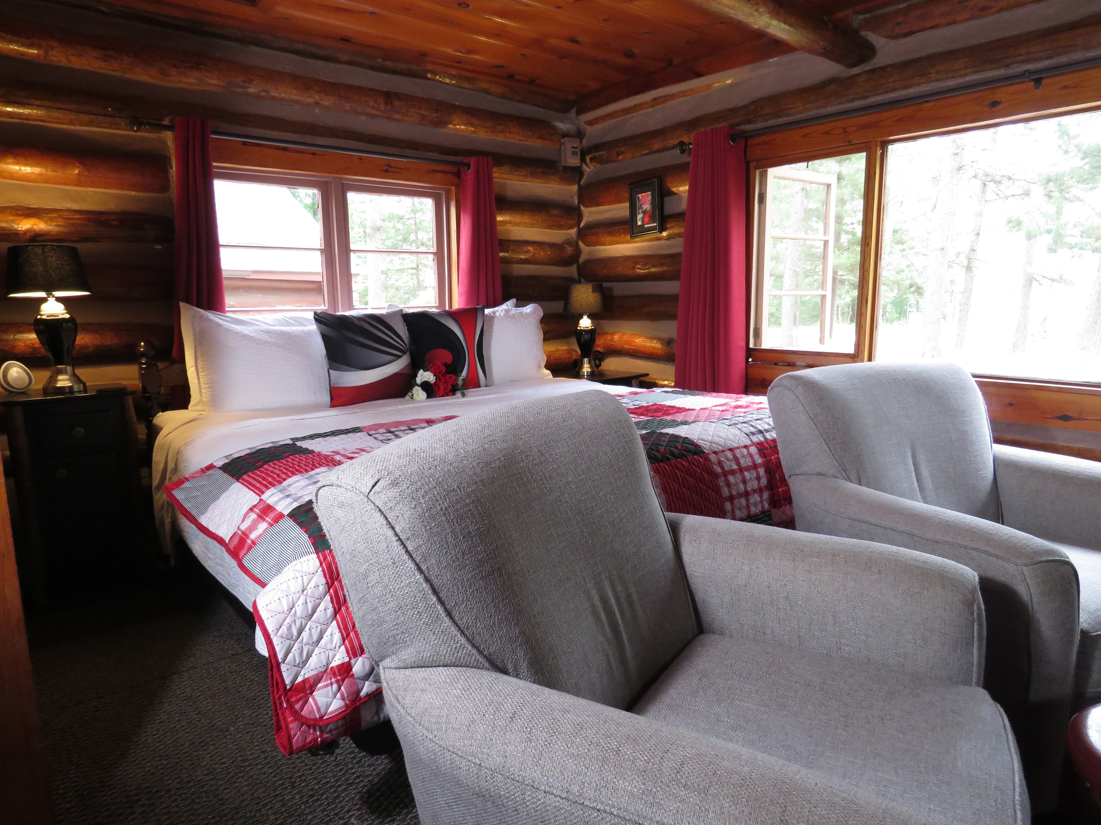 A cozy log cabin bedroom with a king bed, a colorful patchwork quilt, and two upholstered armchairs.