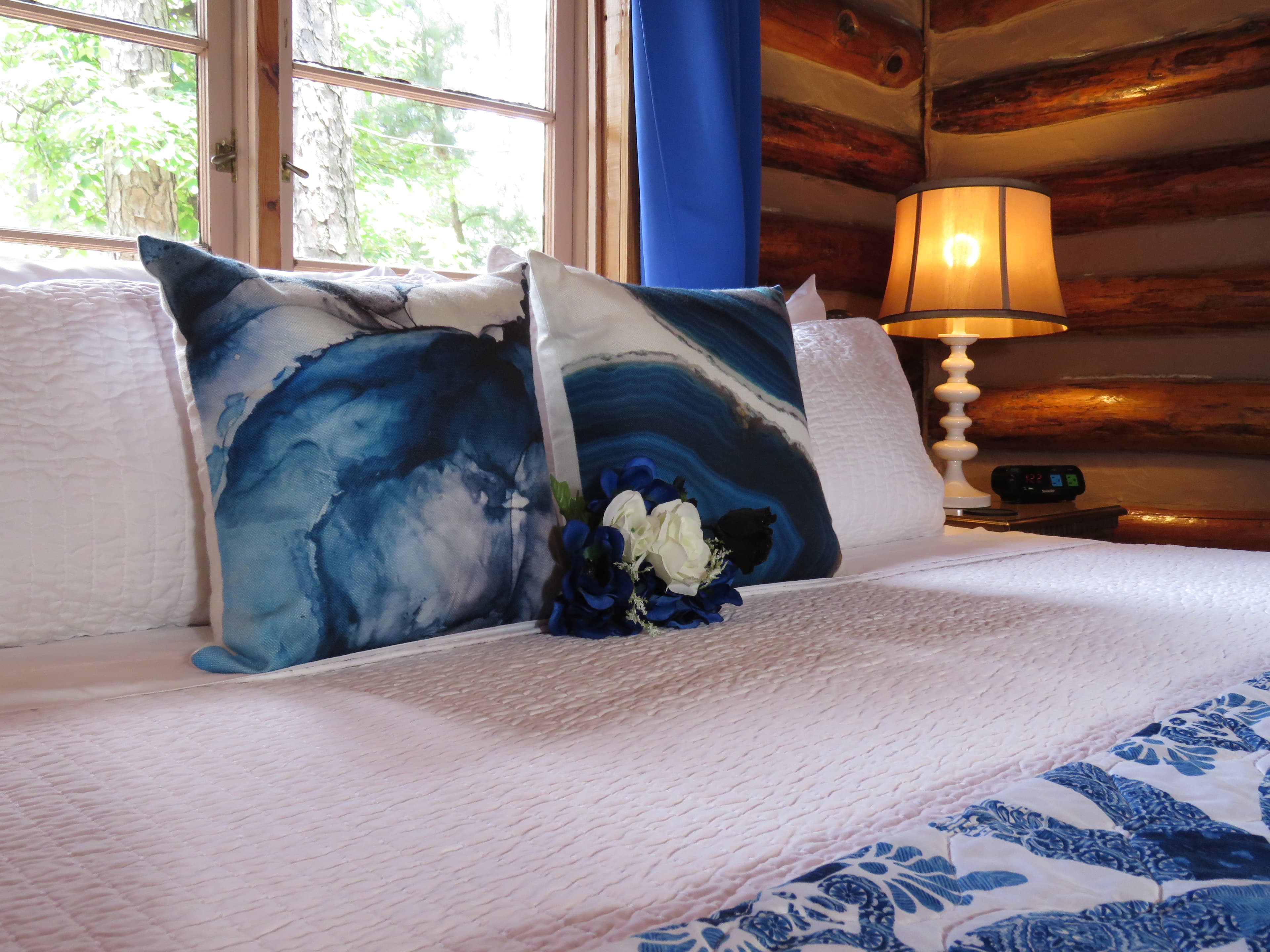 A king-size bed in a log cabin with blue accent pillows, a white comforter, and a bouquet of white and blue flowers as a centerpiece.
