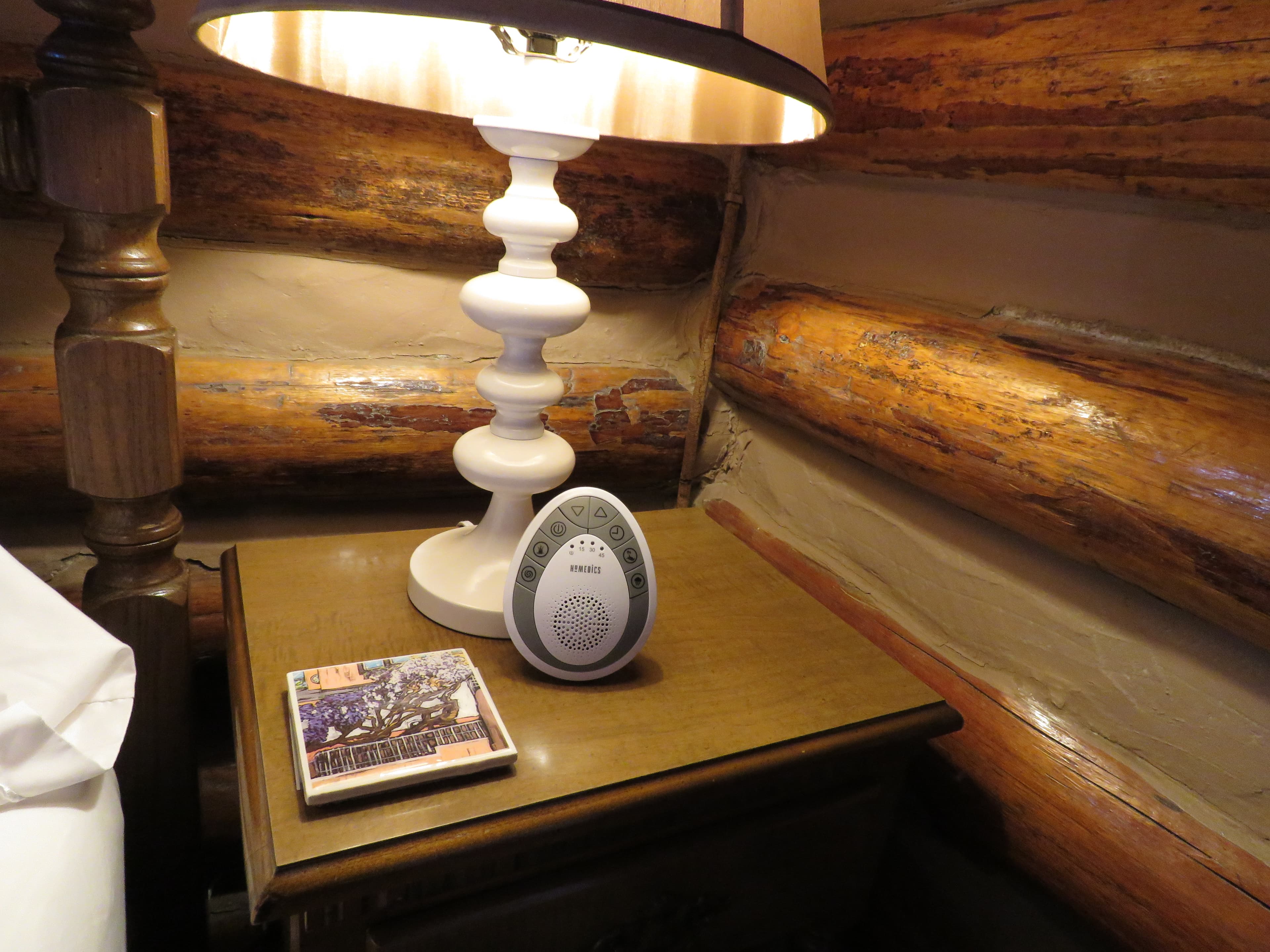 A wooden nightstand holds a white lamp and a white, oval-shaped sound machine. A small book or notepad sits beside them.