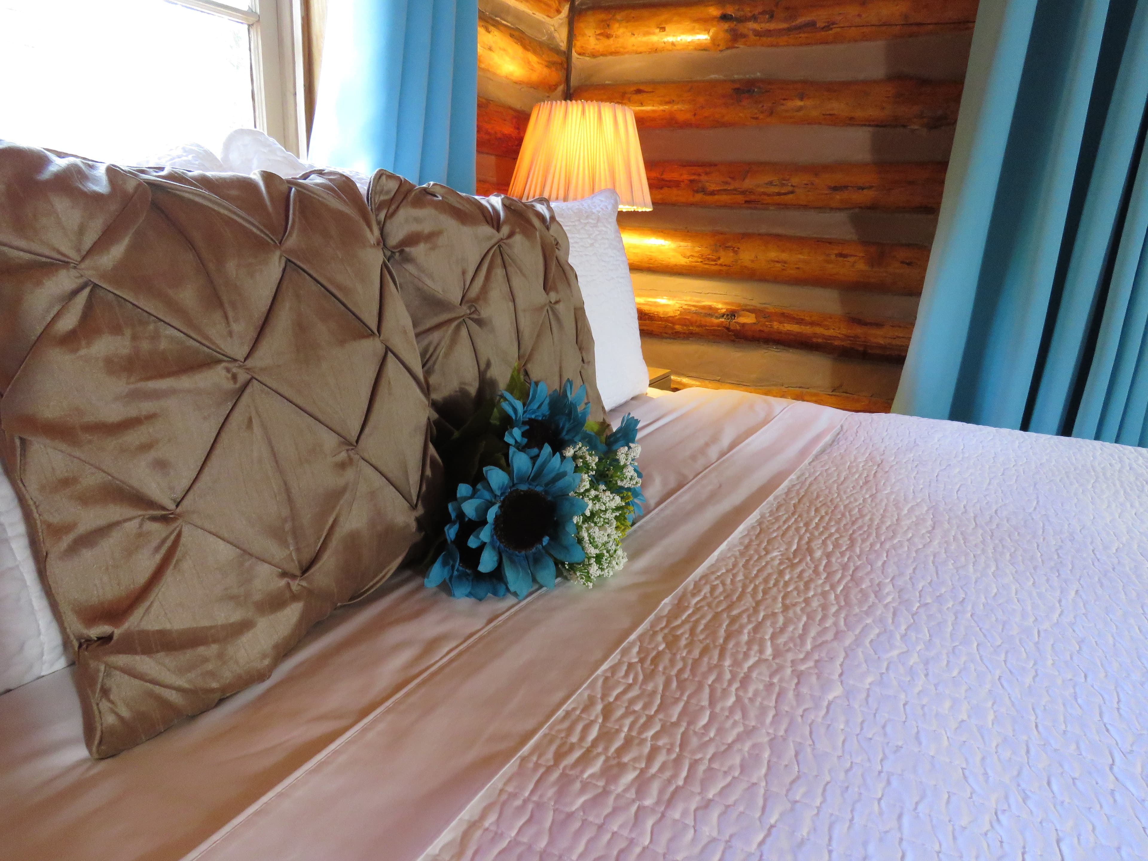 A king bed with a quilted comforter and a decorative gold pillow in a log cabin bedroom. A small bouquet of blue flowers rests on the bed.