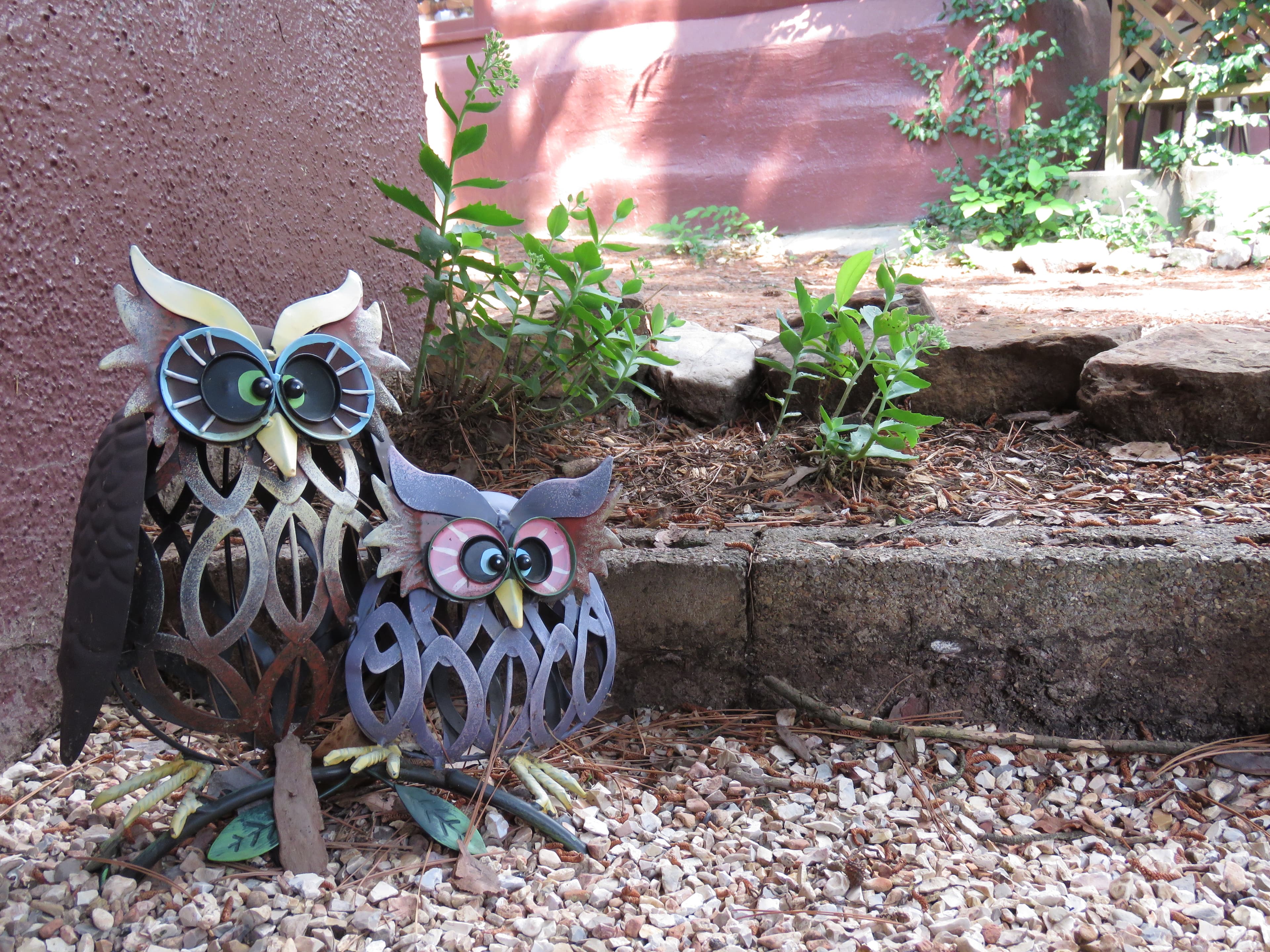Two metallic owl sculptures sit on a bed of gravel. The larger owl is brown and green, while the smaller one is purple.
