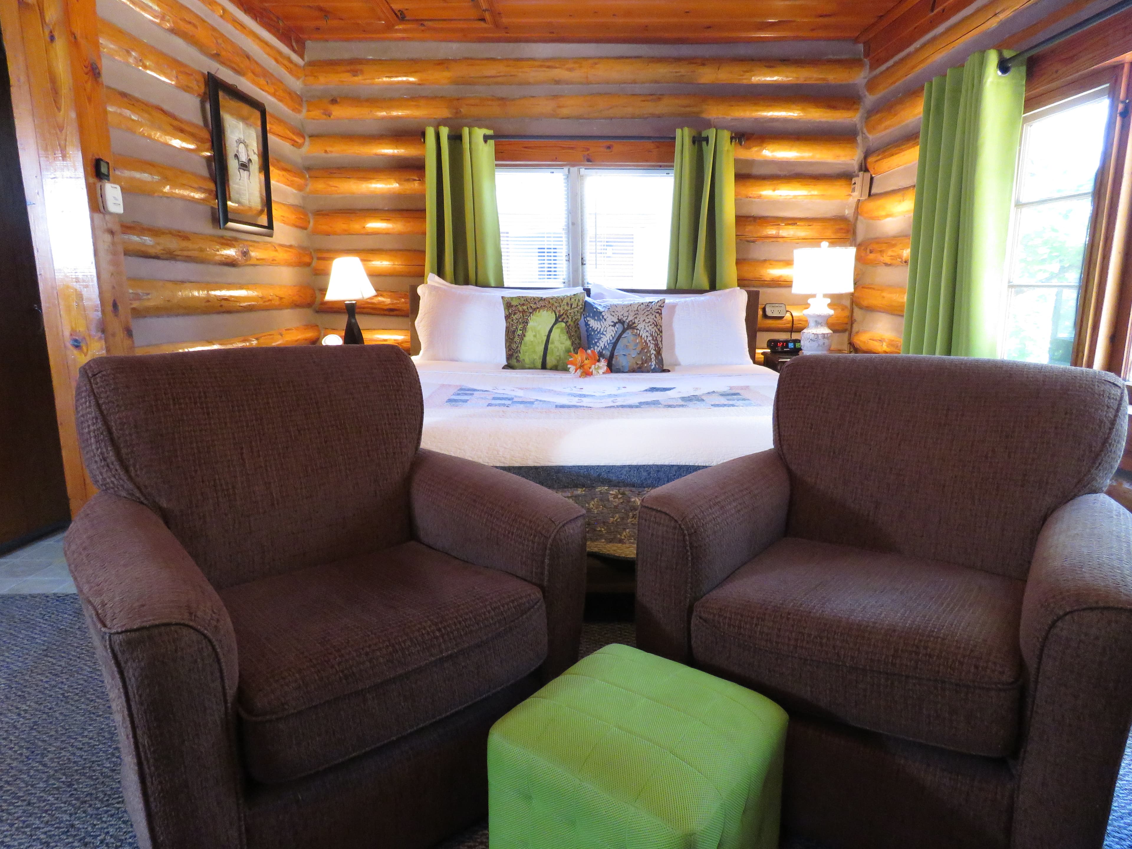 A king-size bed, a pair of armchairs, and a green ottoman are in a cozy log cabin bedroom. The rustic walls and ceiling are made of natural wood.