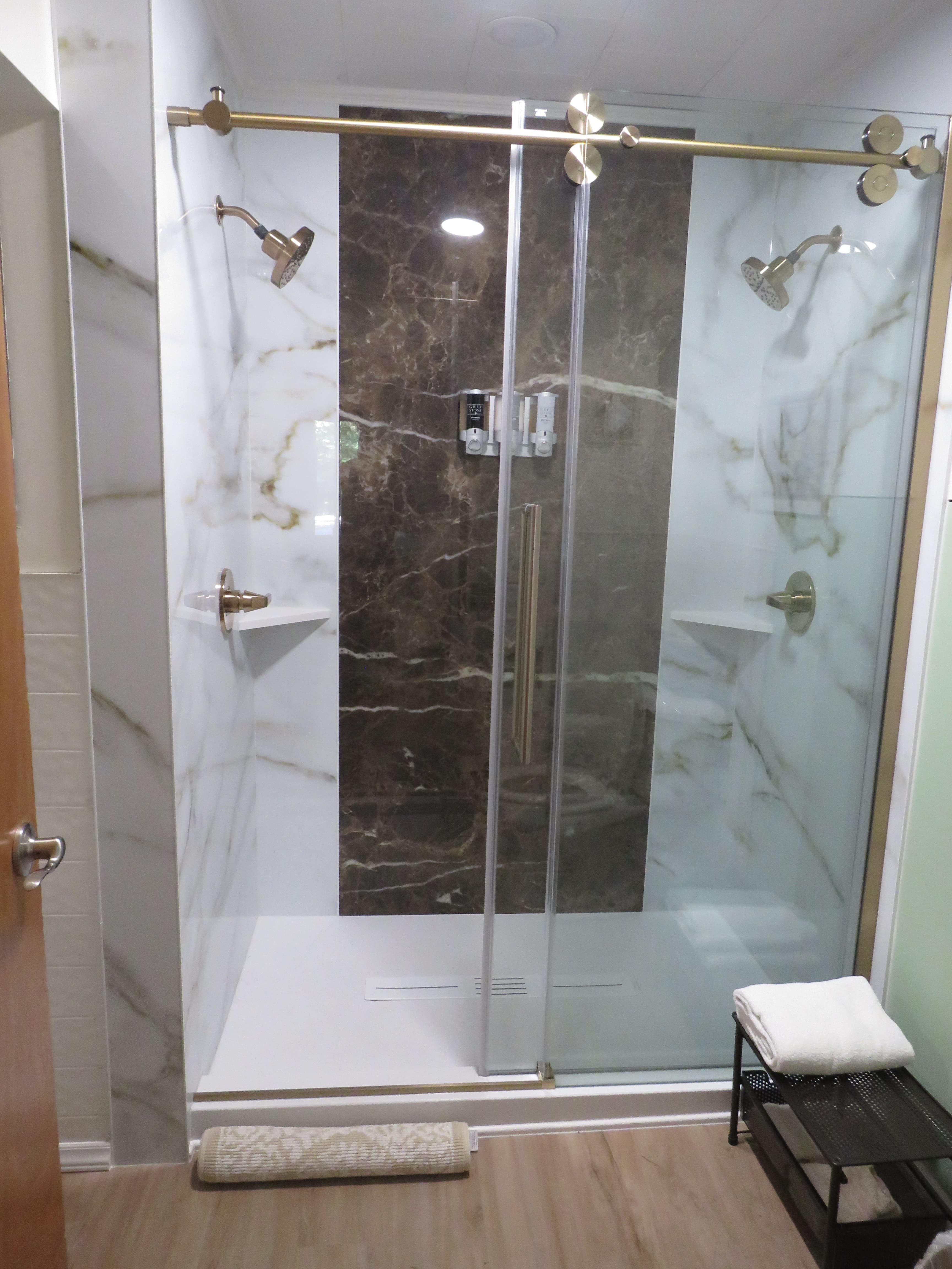 Modern shower with glass doors, dual shower heads, marble walls, and a dark brown accent panel. A small bench with folded towels and a rolled mat are in front of the entrance.
