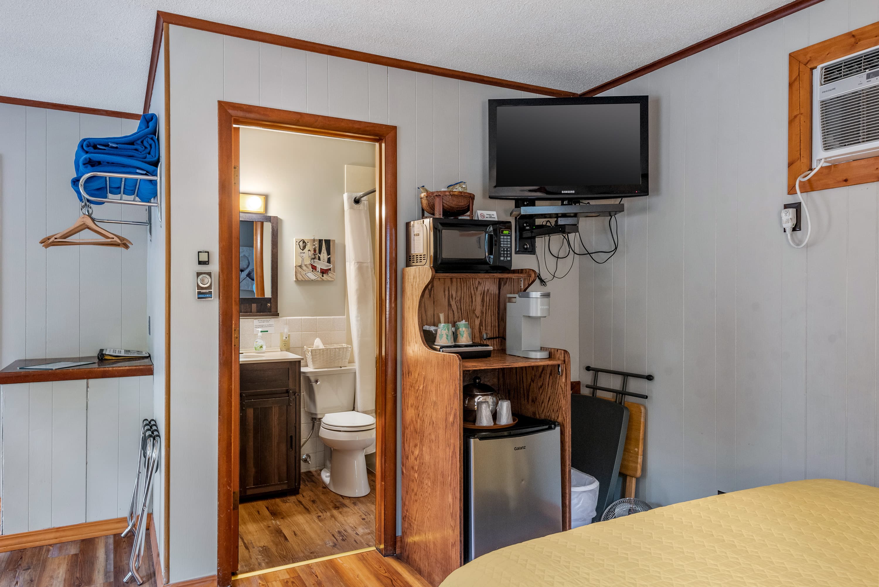 A guest room features a television mounted on a stand with a microwave, a coffee maker, and a mini-fridge. An open doorway leads to a bathroom.
