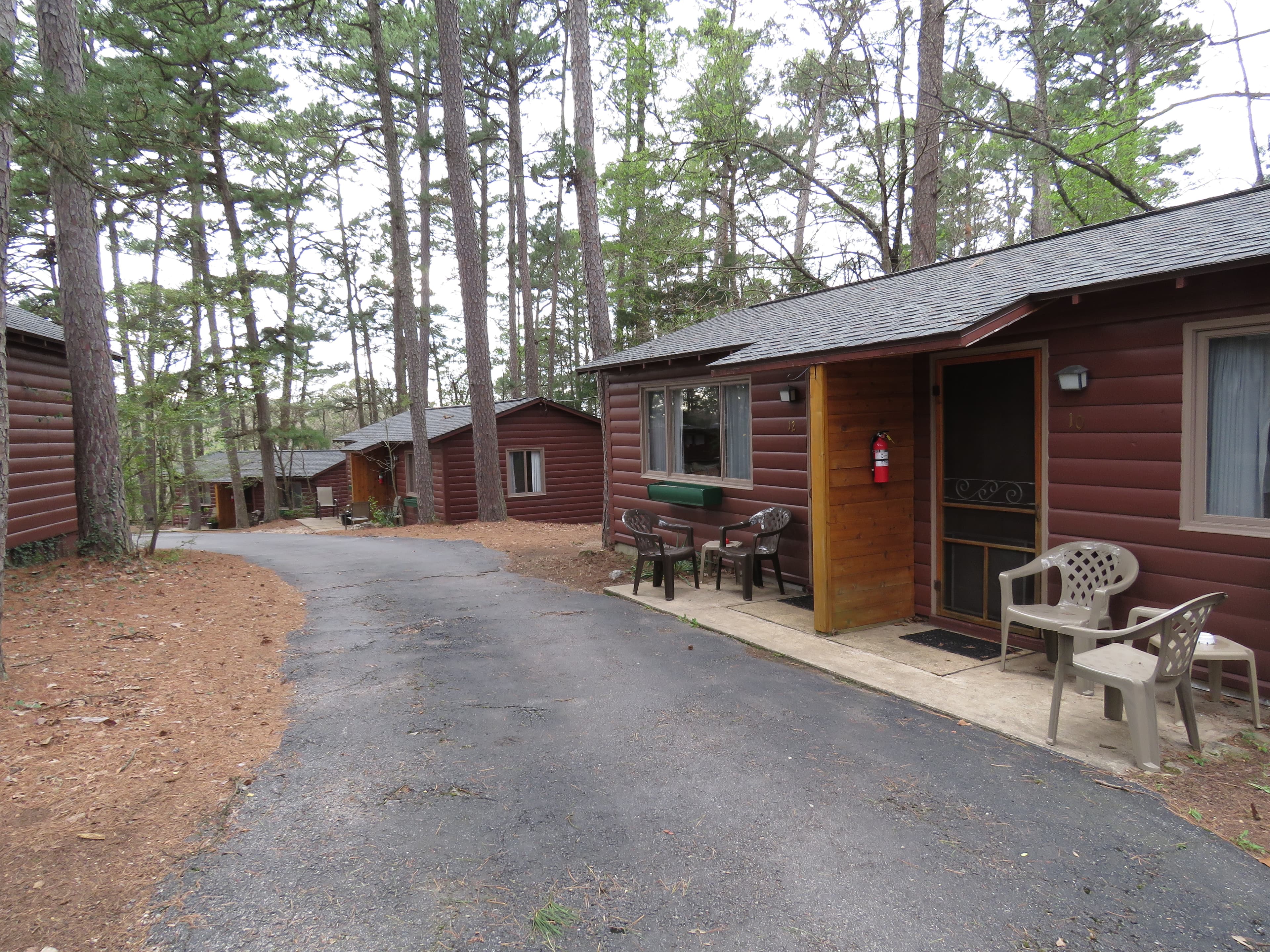A paved driveway winds past several rustic log cabins, each with a small covered porch and chairs. The cabins are nestled in a wooded area.