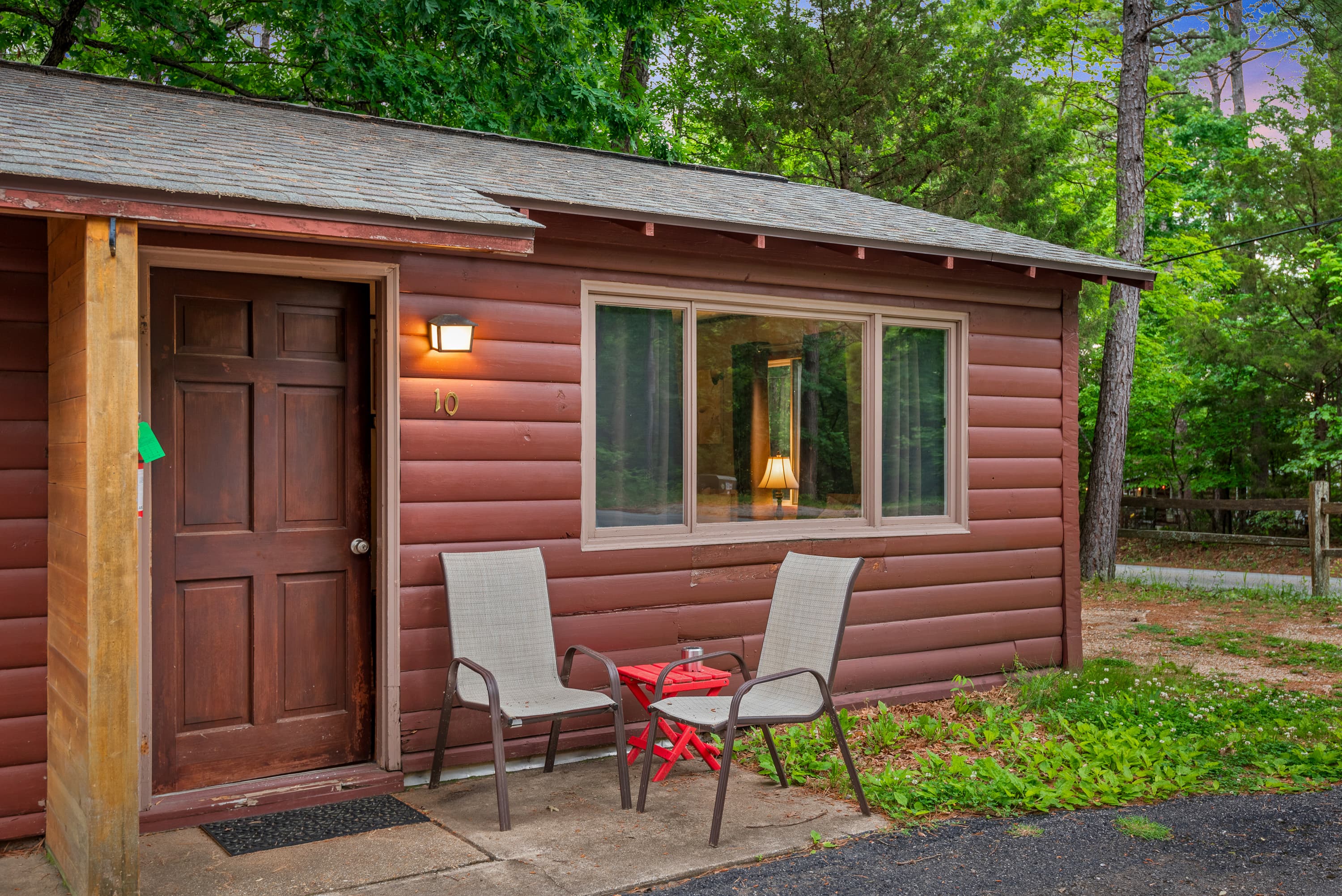 A charming, rustic cabin with a covered porch holding two chairs and a small red table. The cabin is nestled in a wooded area.