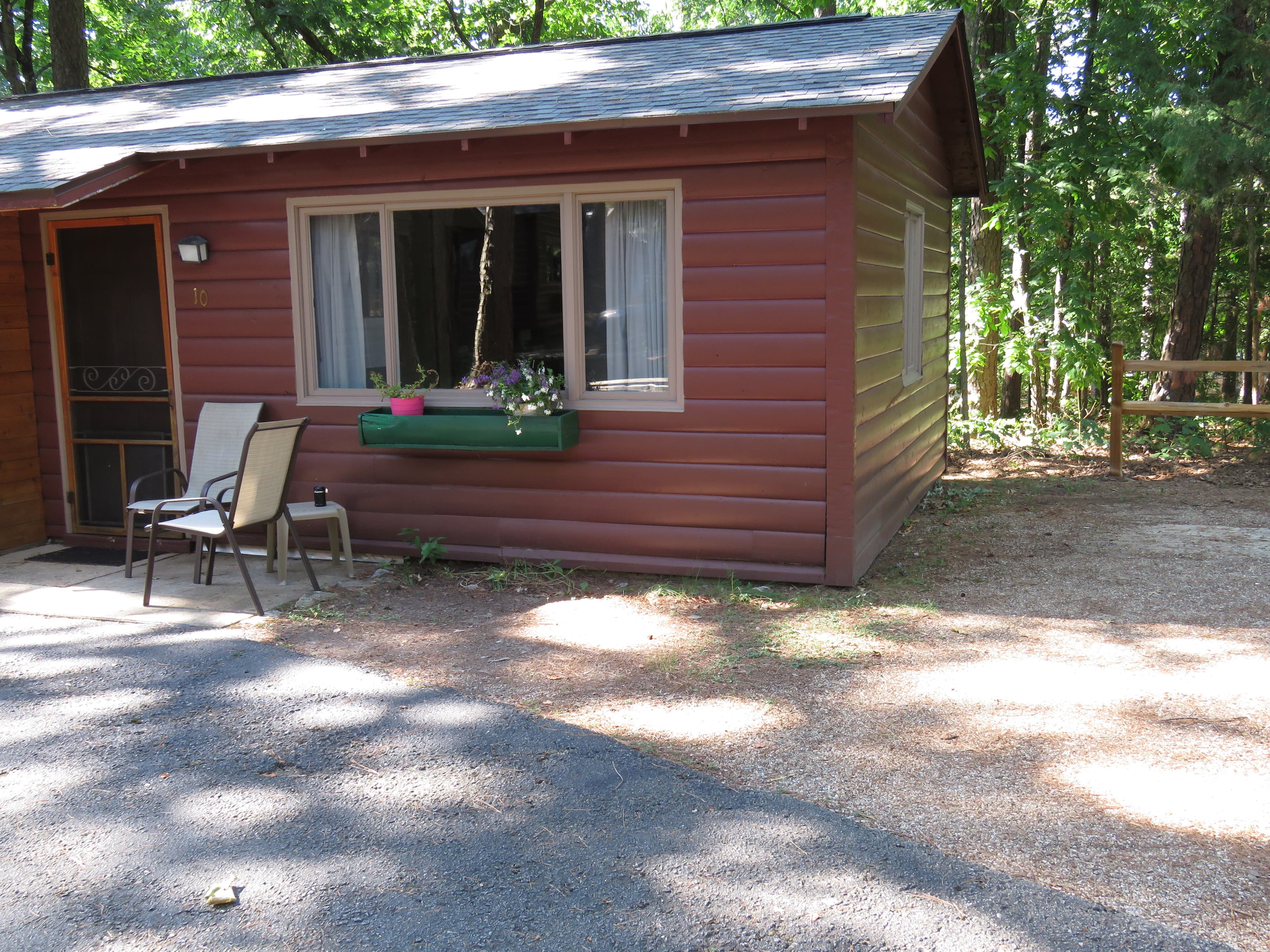 A cozy, small cabin with a front window that has a flower box and a small porch with two chairs. The cabin is surrounded by a wooded area.