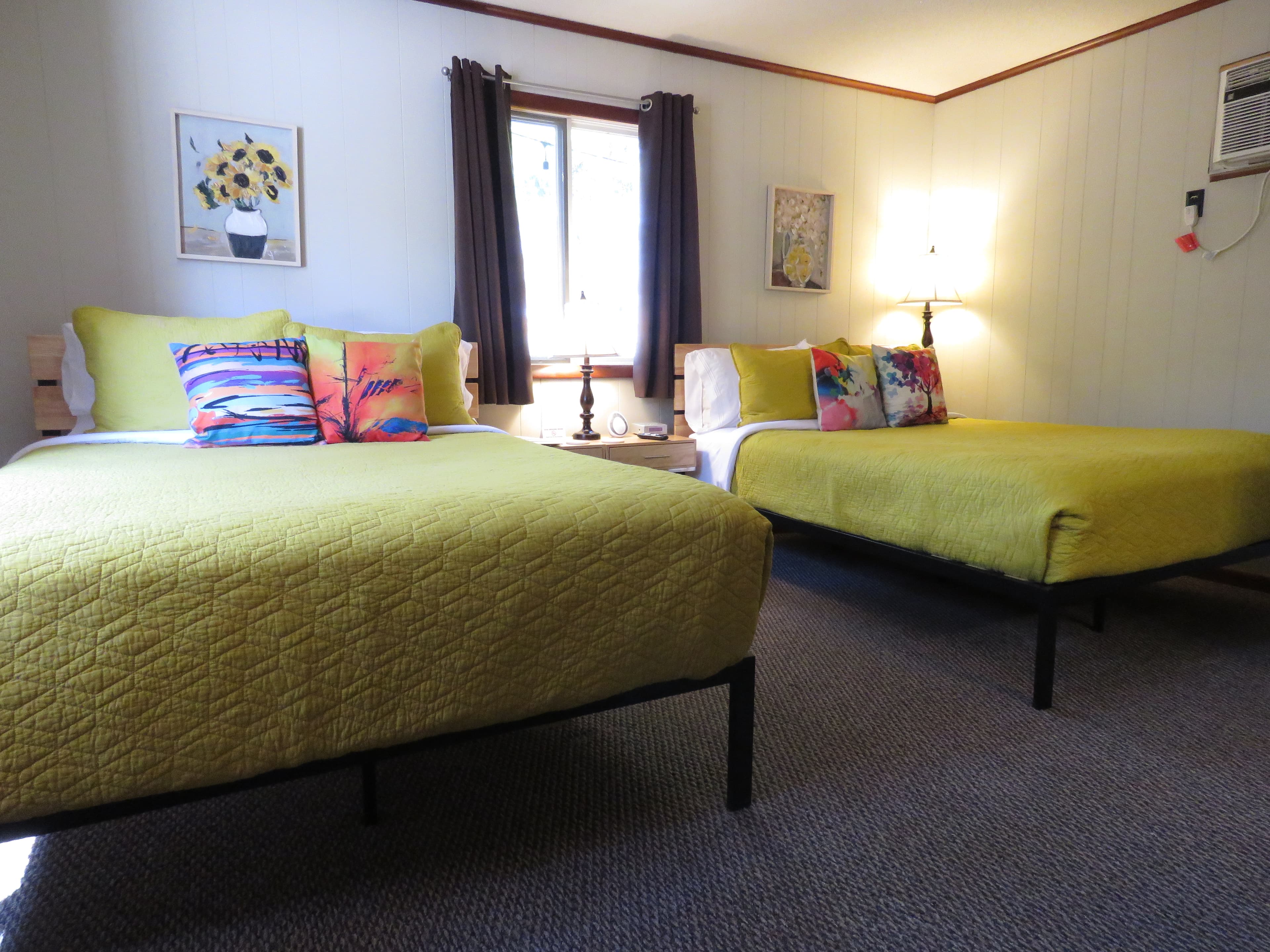 A bedroom features two queen beds with bright yellow quilts and colorful pillows. The walls are a neutral color, and there are two framed pictures on the wall.