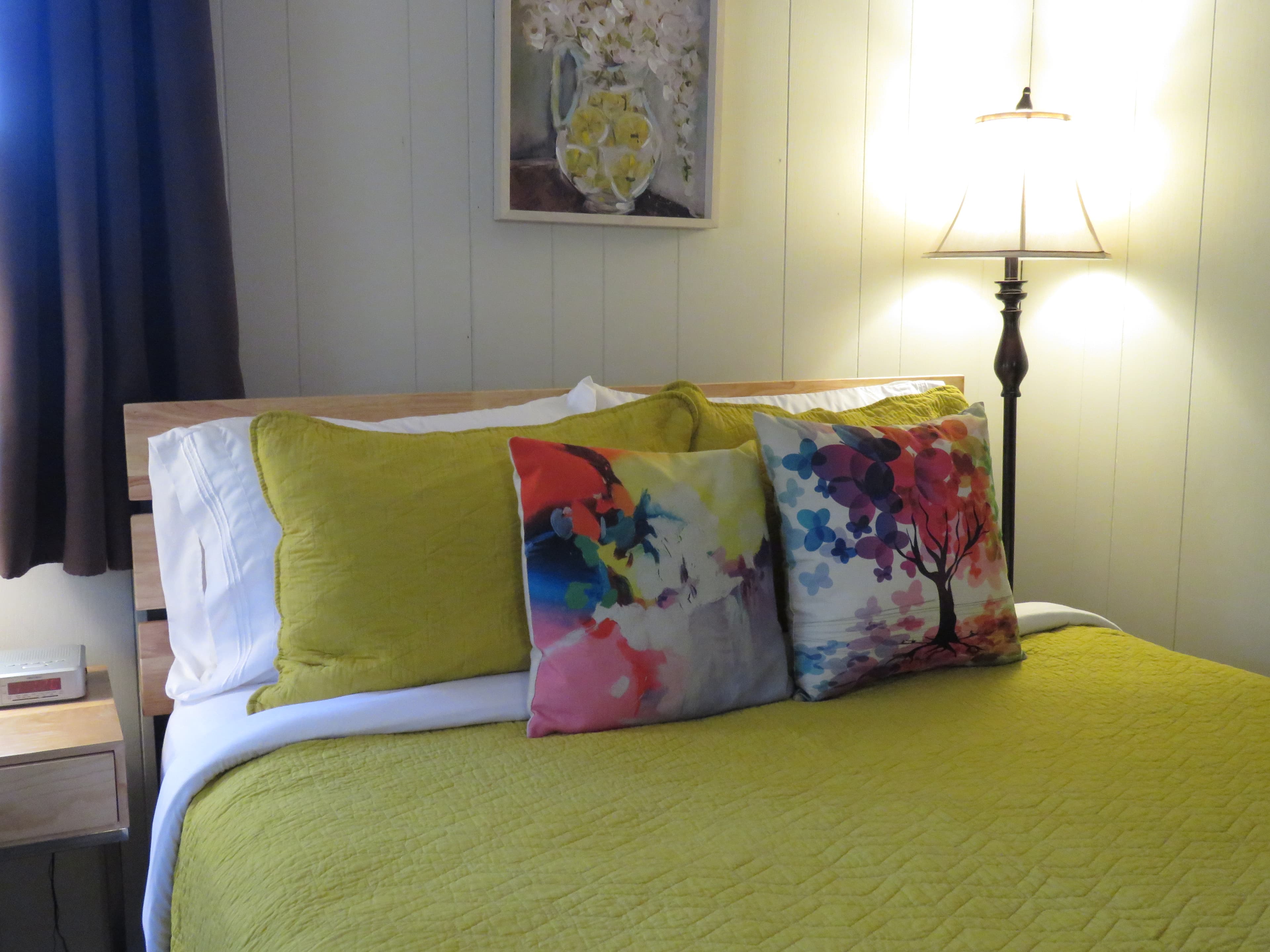 A queen bed with a bright yellow comforter and three pillows. The pillows include a solid yellow one and two with colorful, modern patterns.
