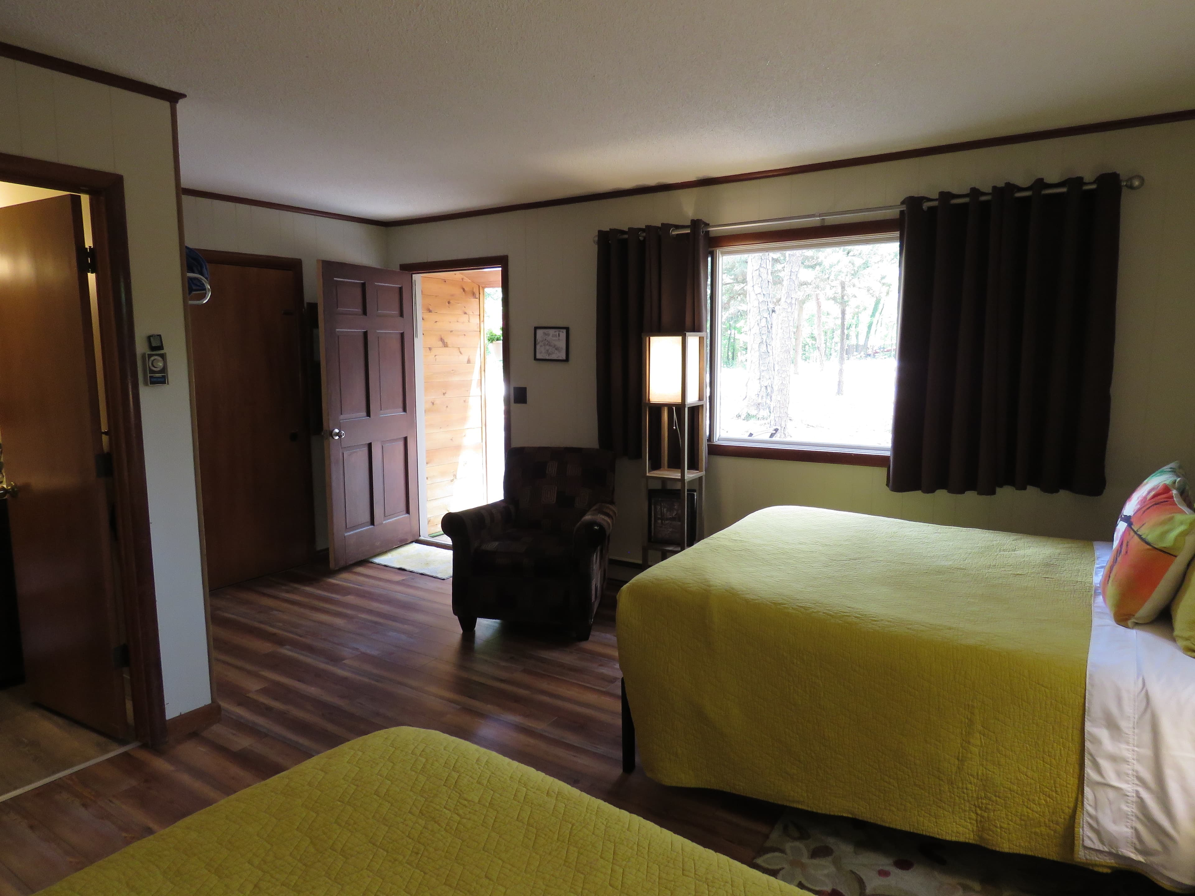 A cozy room features two queen beds with bright yellow quilts, a comfortable armchair, and a small table. A doorway leads to the outside.