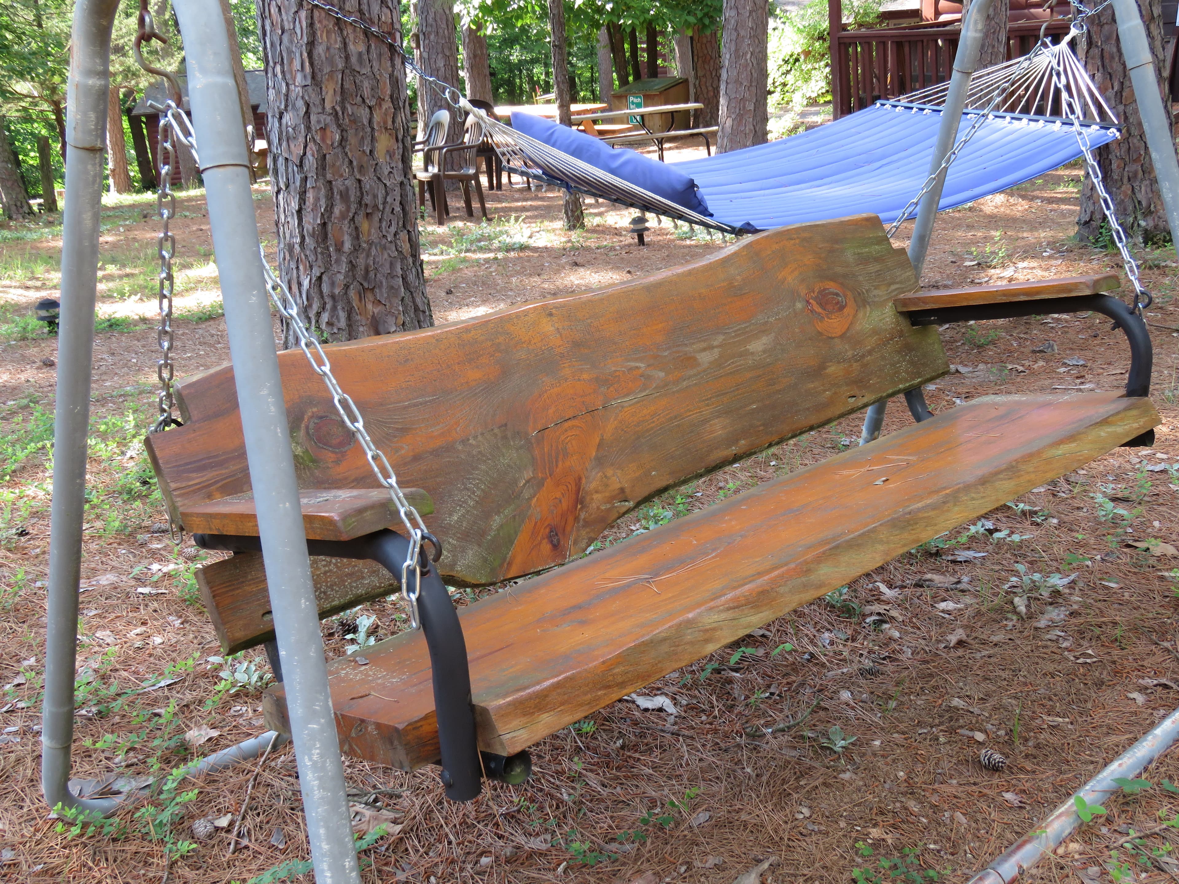 A rustic wooden bench swing with a natural-edge back is shown in a wooded area. A blue hammock is visible in the background.