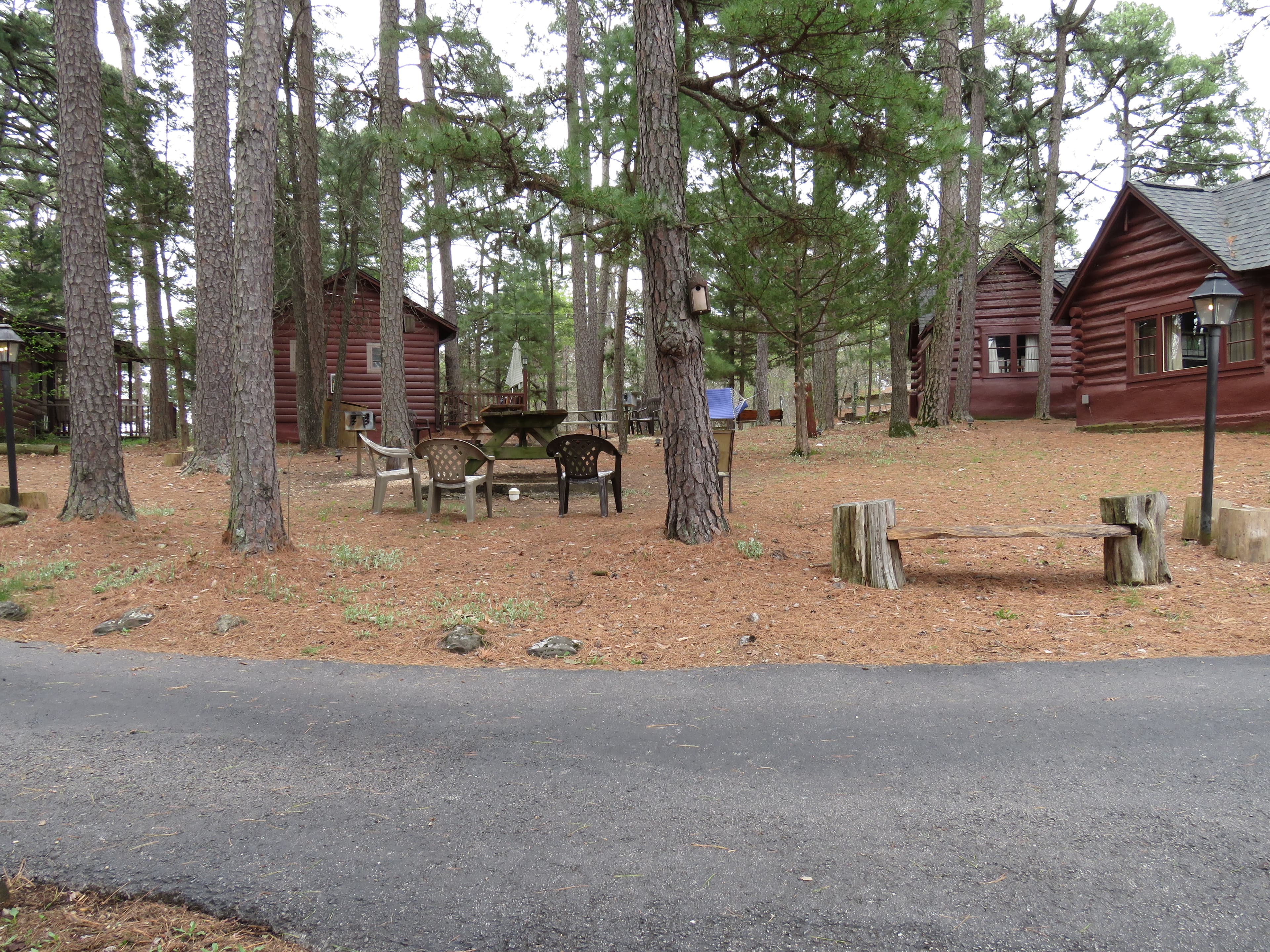 A view of a wooded area with several rustic cabins. The foreground features a seating area with four chairs, a bench, and a gravel road.