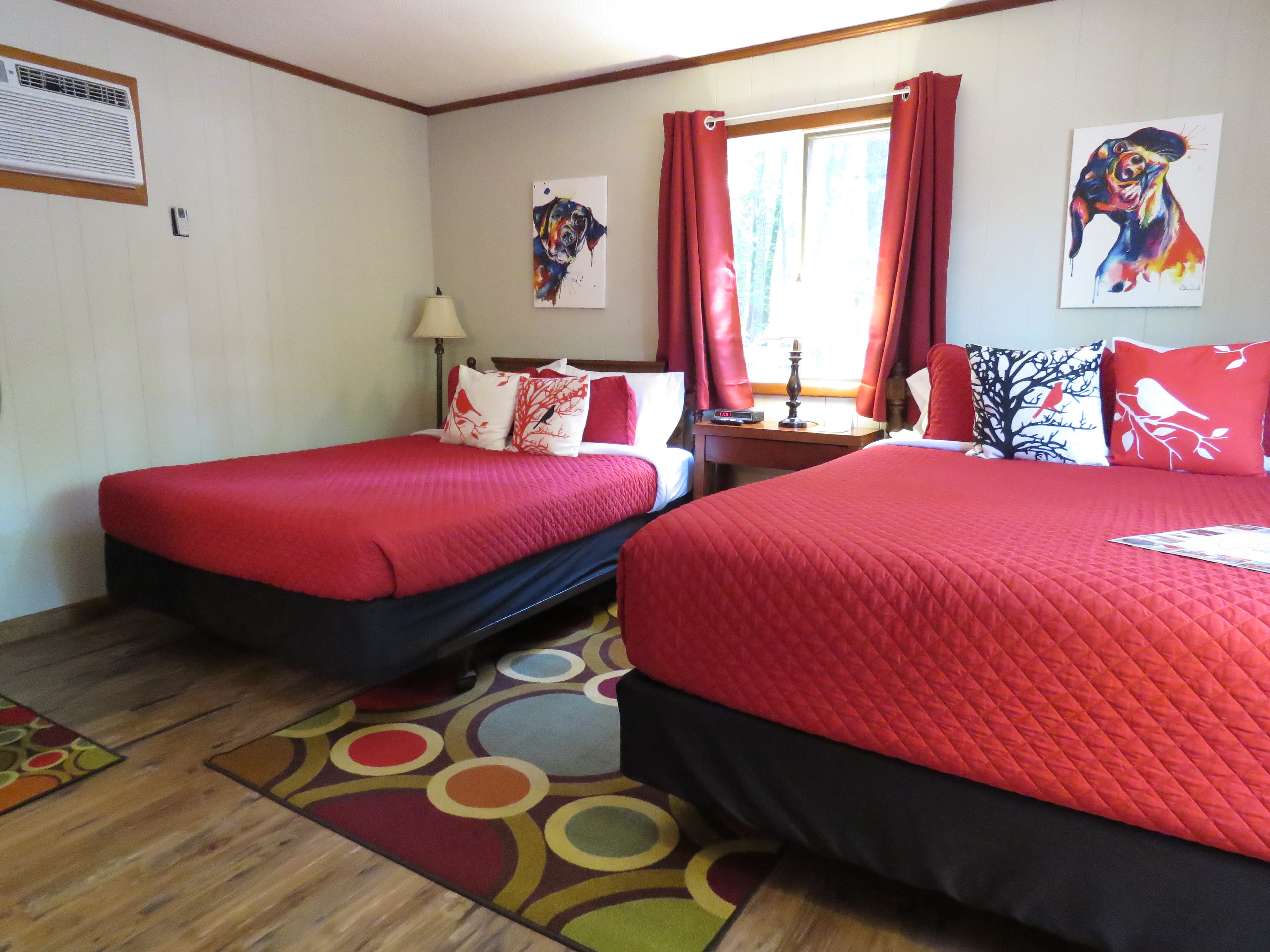 A cozy room features two beds with bright red quilts and dog-themed art on the walls. A colorful, patterned rug is on the floor.