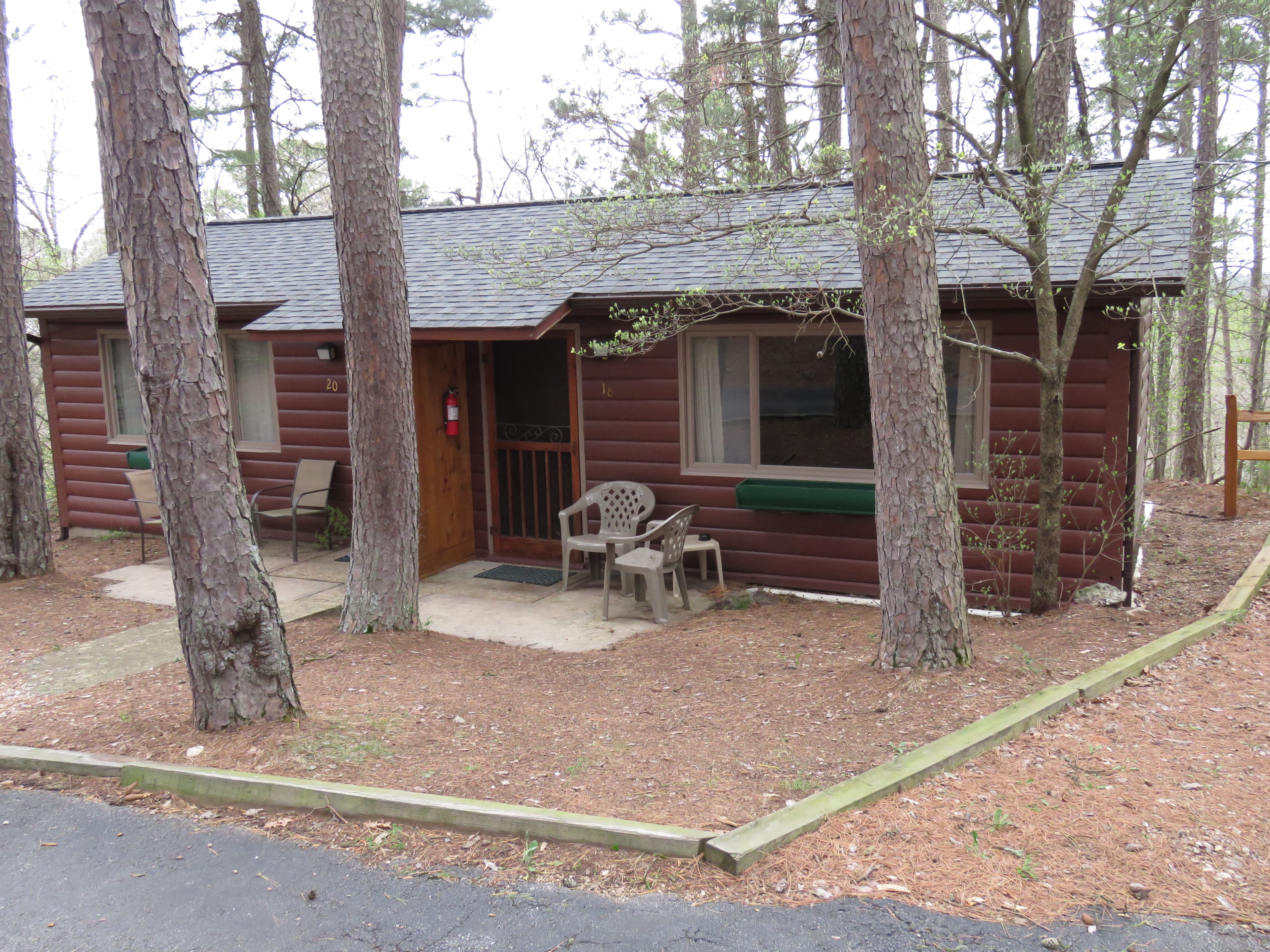A charming, rustic duplex cabin with a paved driveway. Each unit has a small porch with a table and chairs, and there are windows with flower boxes.