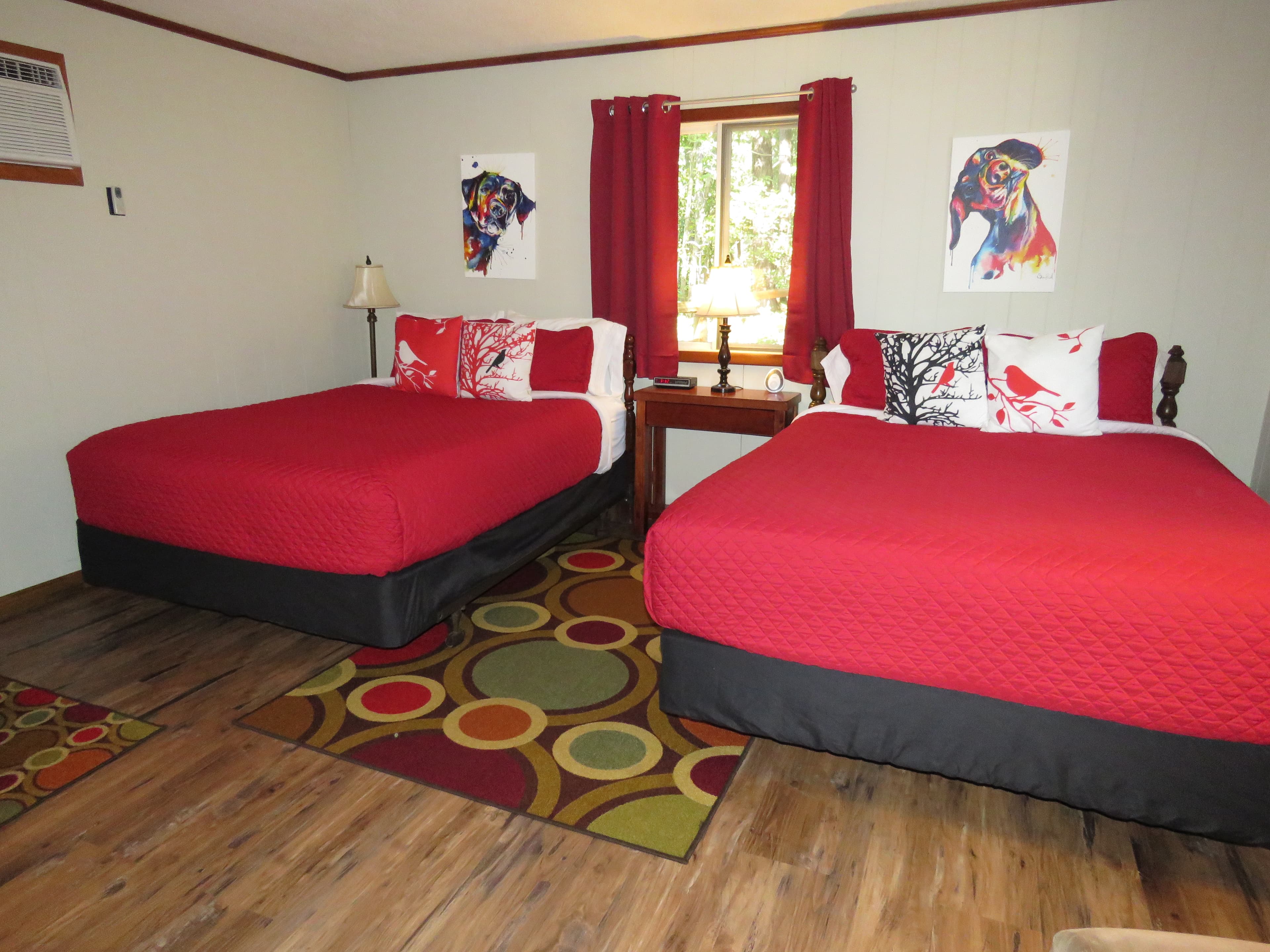 A cozy room features two beds with bright red quilts and dog-themed art on the walls. A colorful, patterned rug is on the floor.
