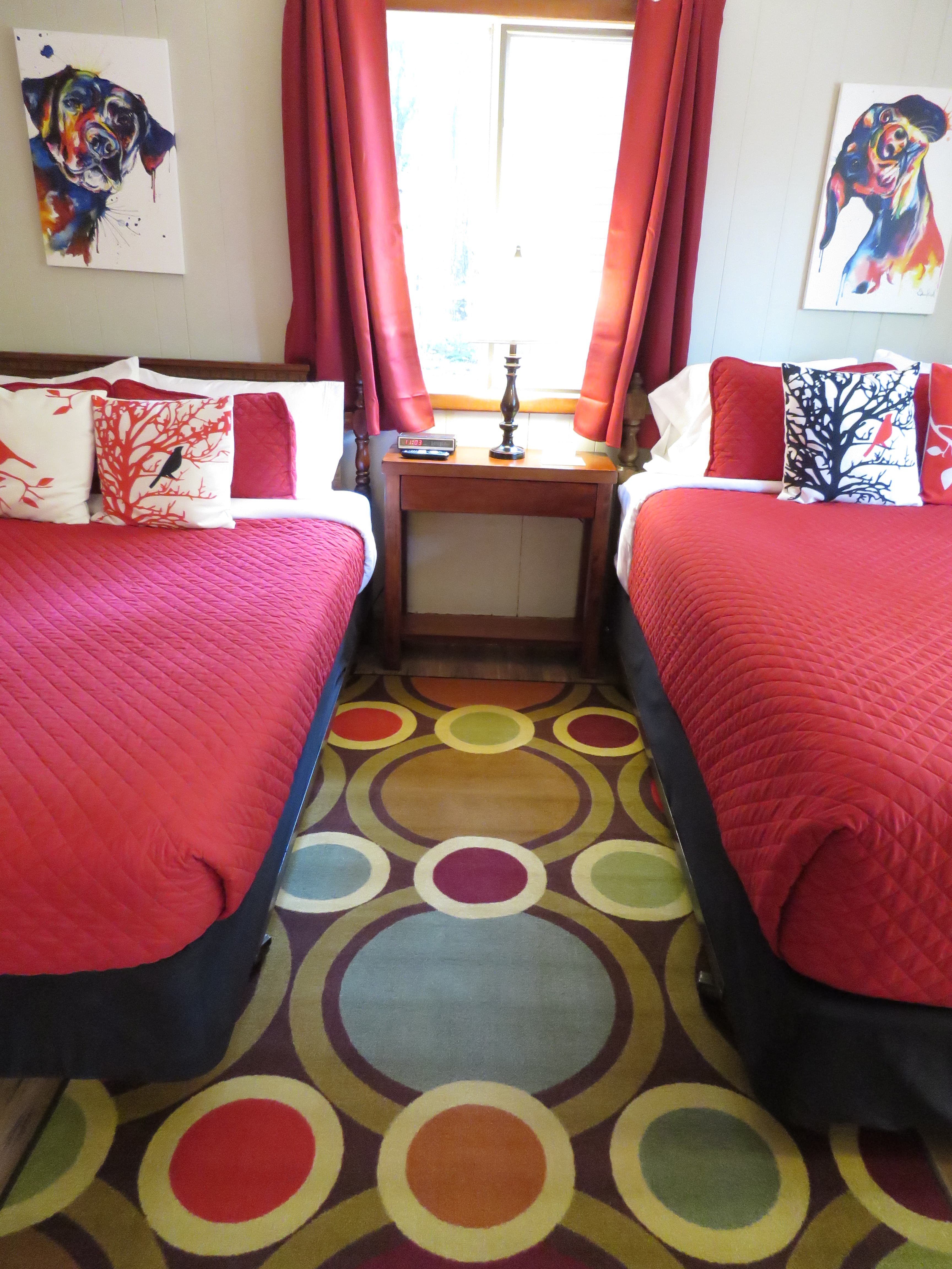A cozy room features two beds with bright red quilts and dog-themed art on the walls. A colorful, patterned rug is on the floor.