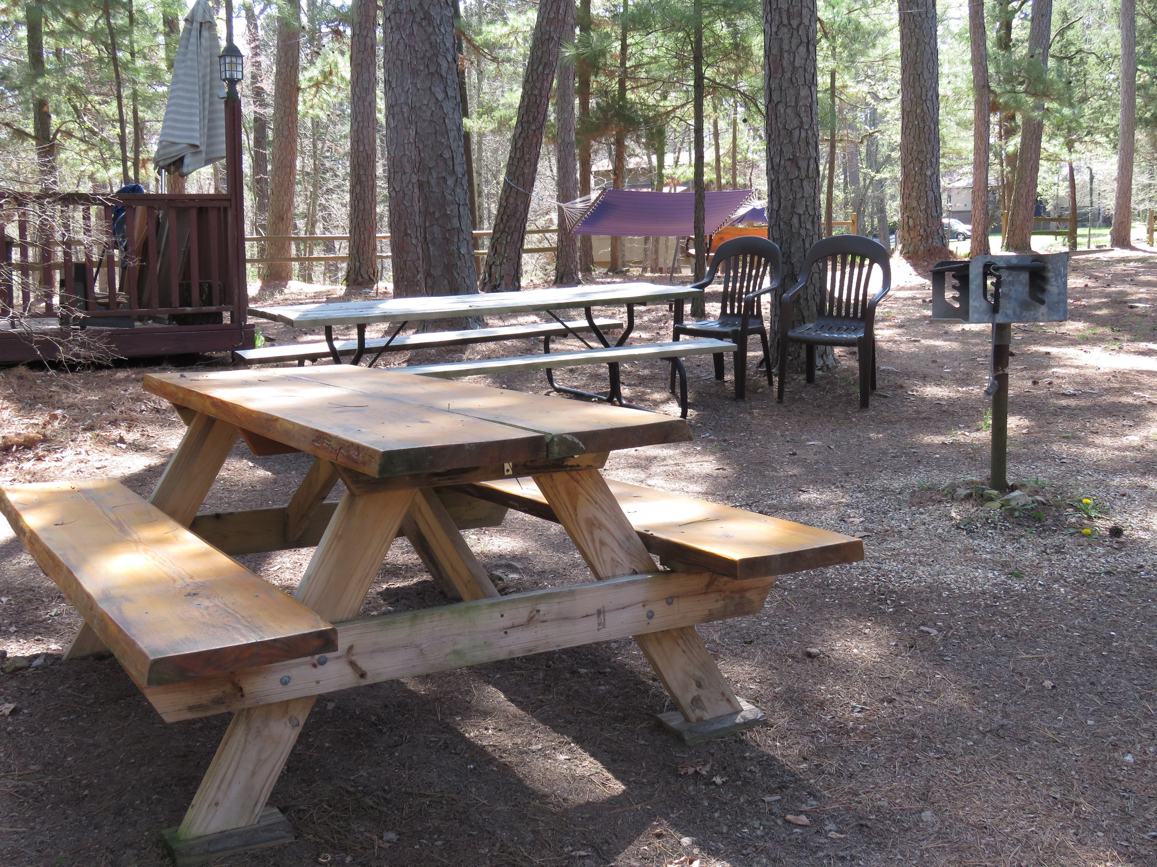 A rustic outdoor area features two wooden picnic tables and a grill. In the background, there is a deck with an umbrella and chairs.