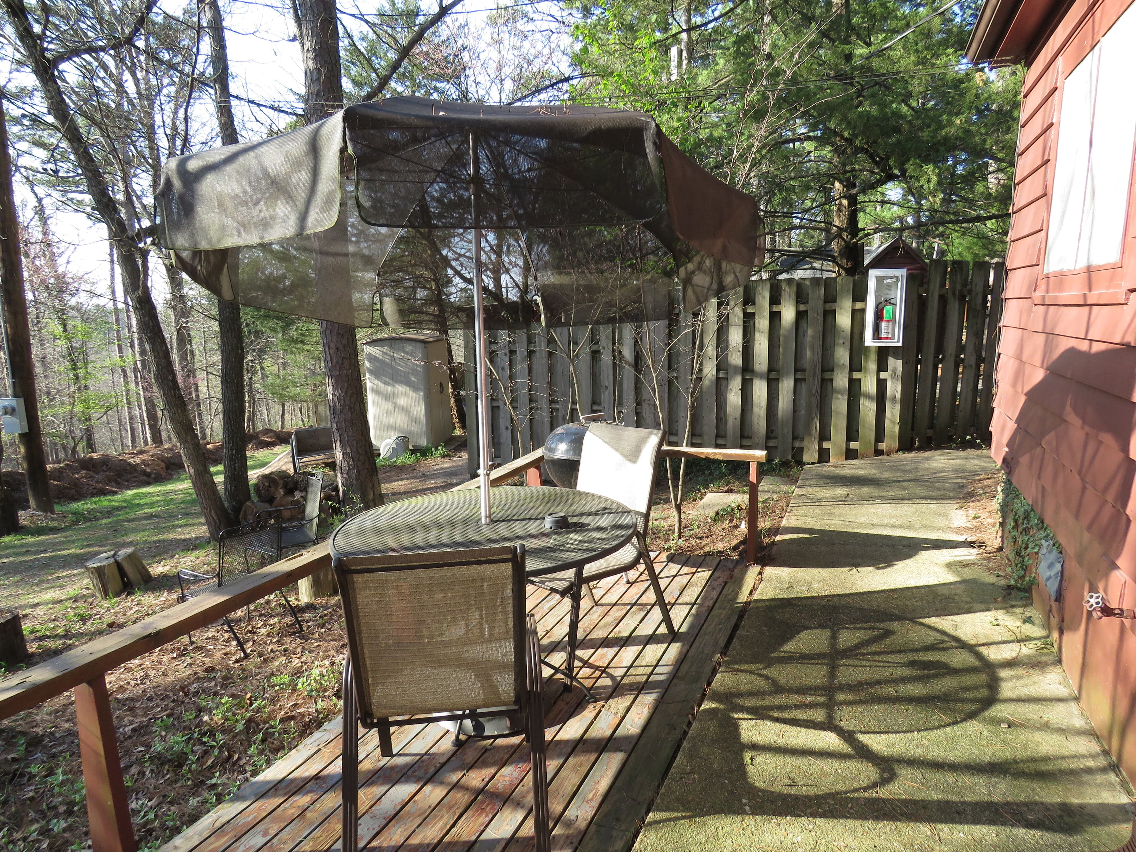 A wooden deck features a round table, two chairs, and a black table umbrella. A brown picket fence is in the background.