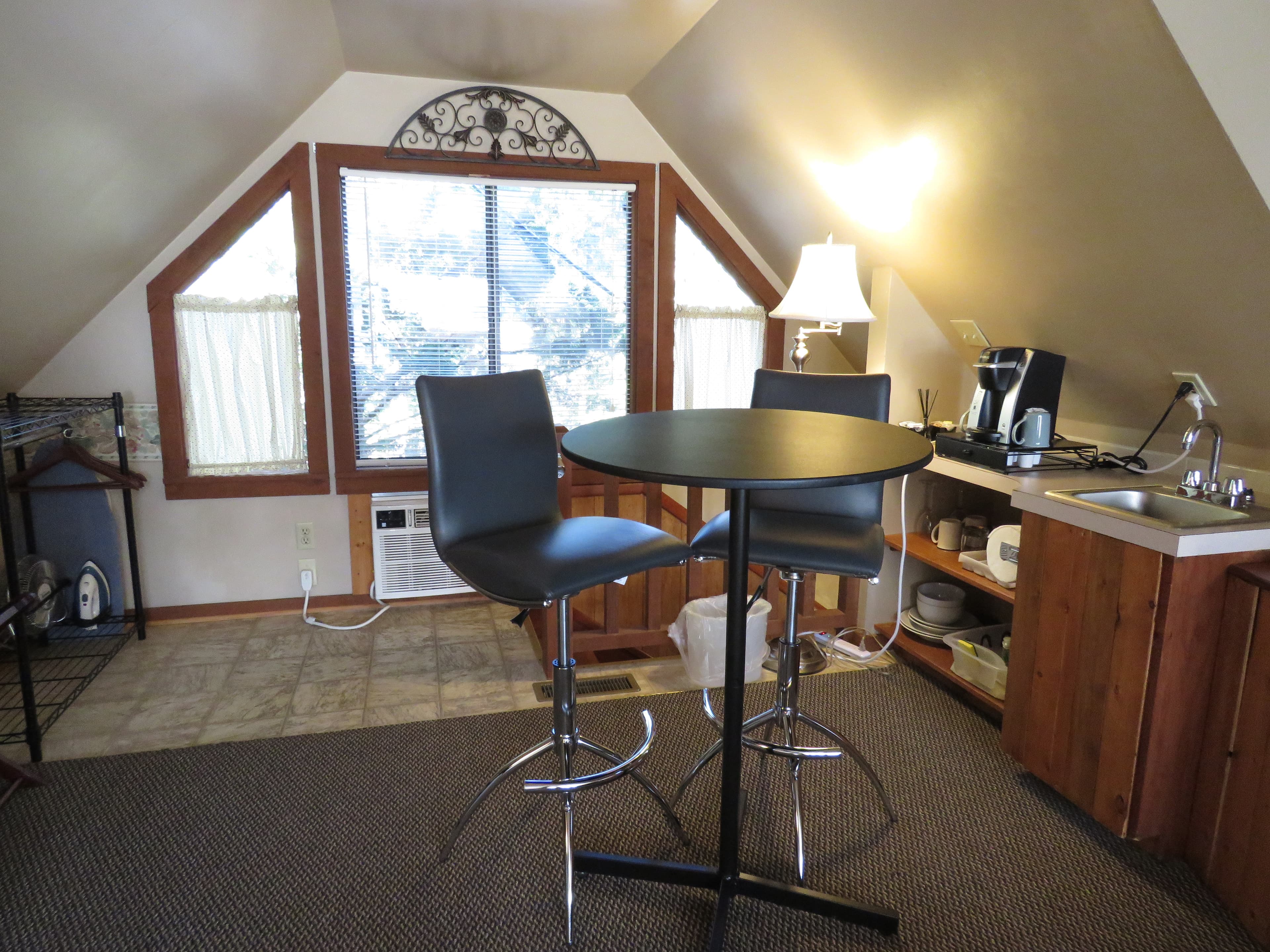 A loft area features a small round table with two barstools, a mini-kitchenette, and a large windows at the far end of the room.