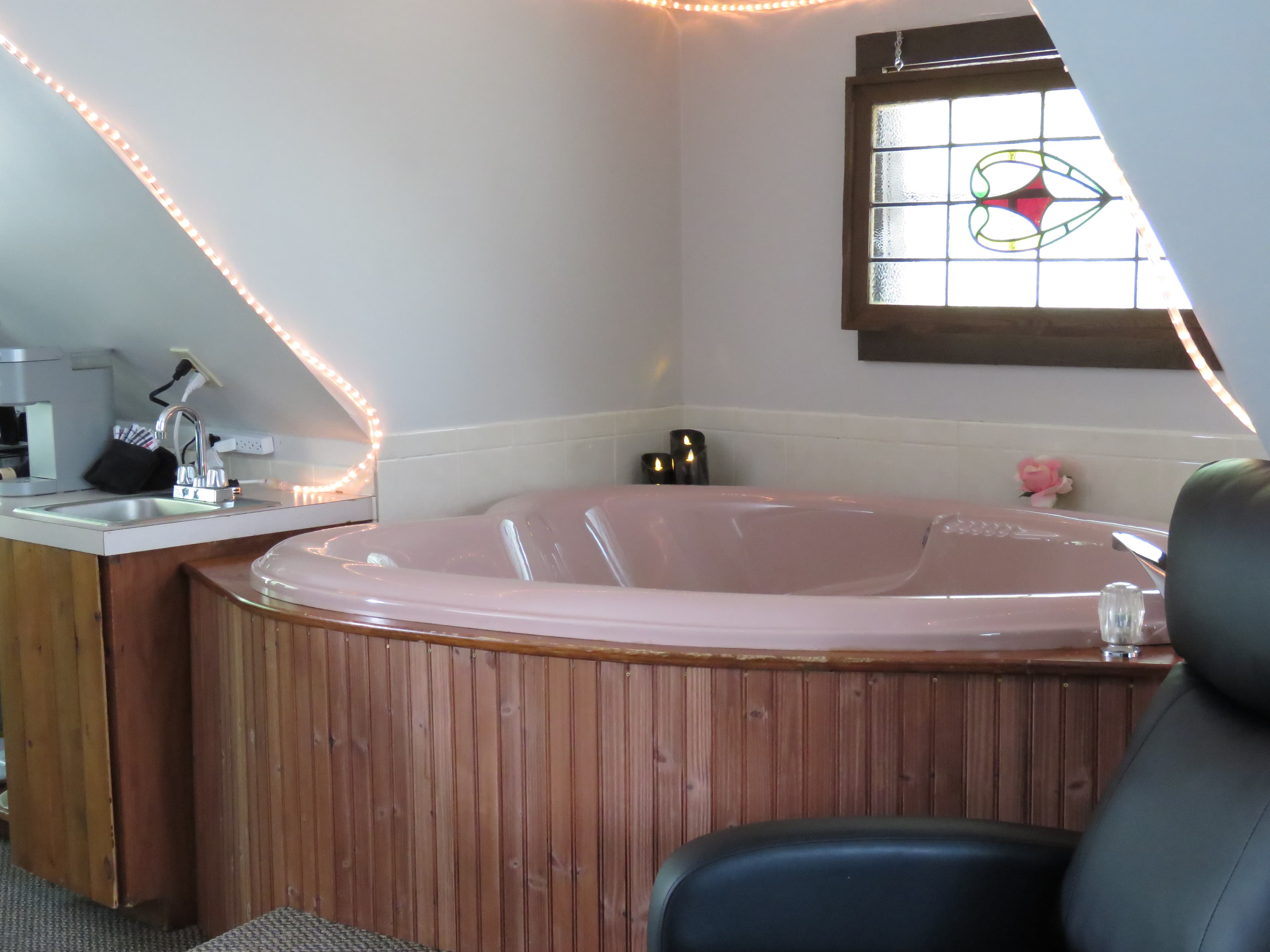 A cozy loft with a large, pink jetted tub. A stained-glass window is on the wall, and string lights are strung on the sloped ceiling.