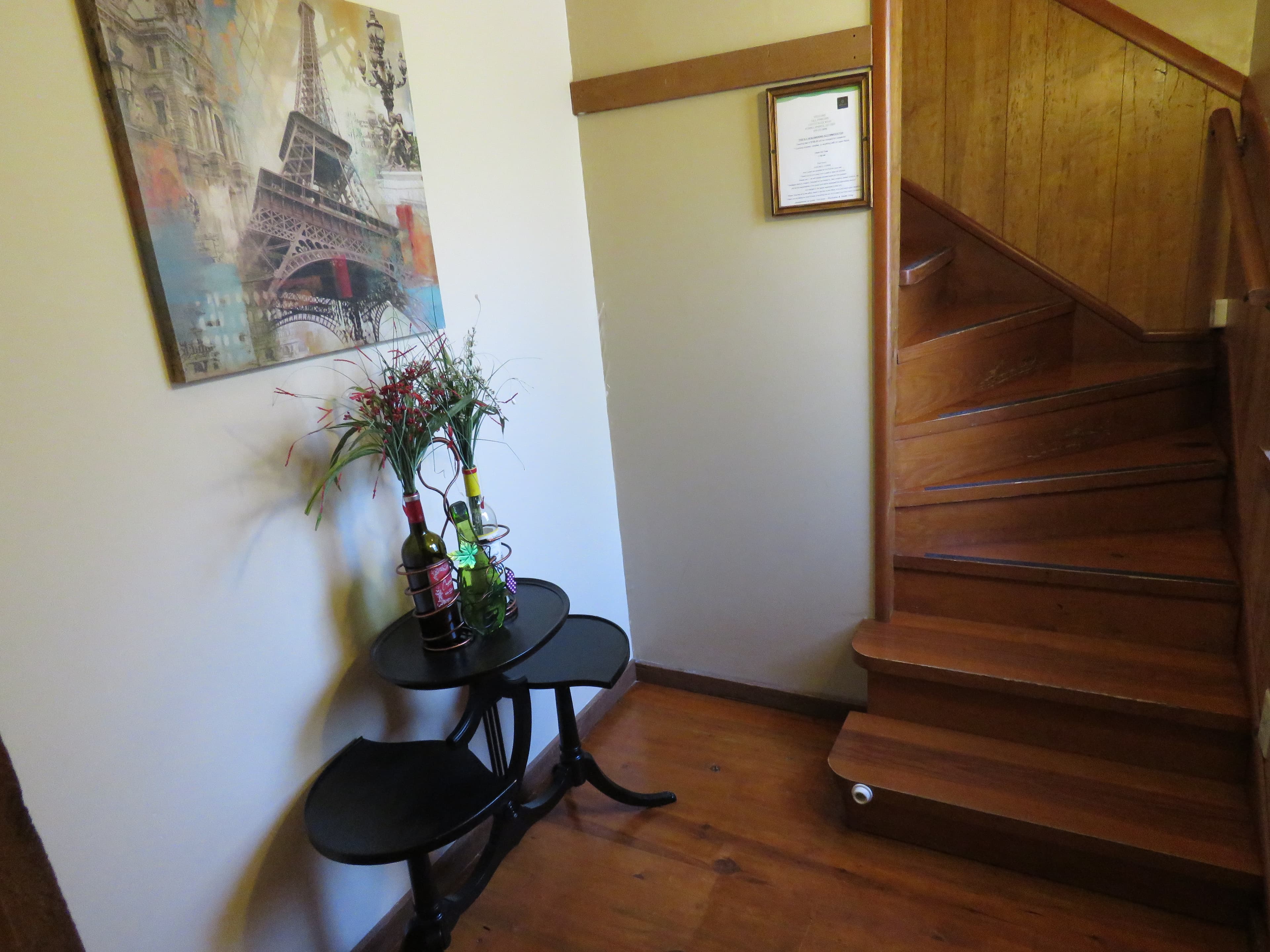 A rustic staircase with wooden steps and banisters is on the right. On the left, a small table holds two bottles of wine, and a framed picture of the Eiffel Tower is on the wall.