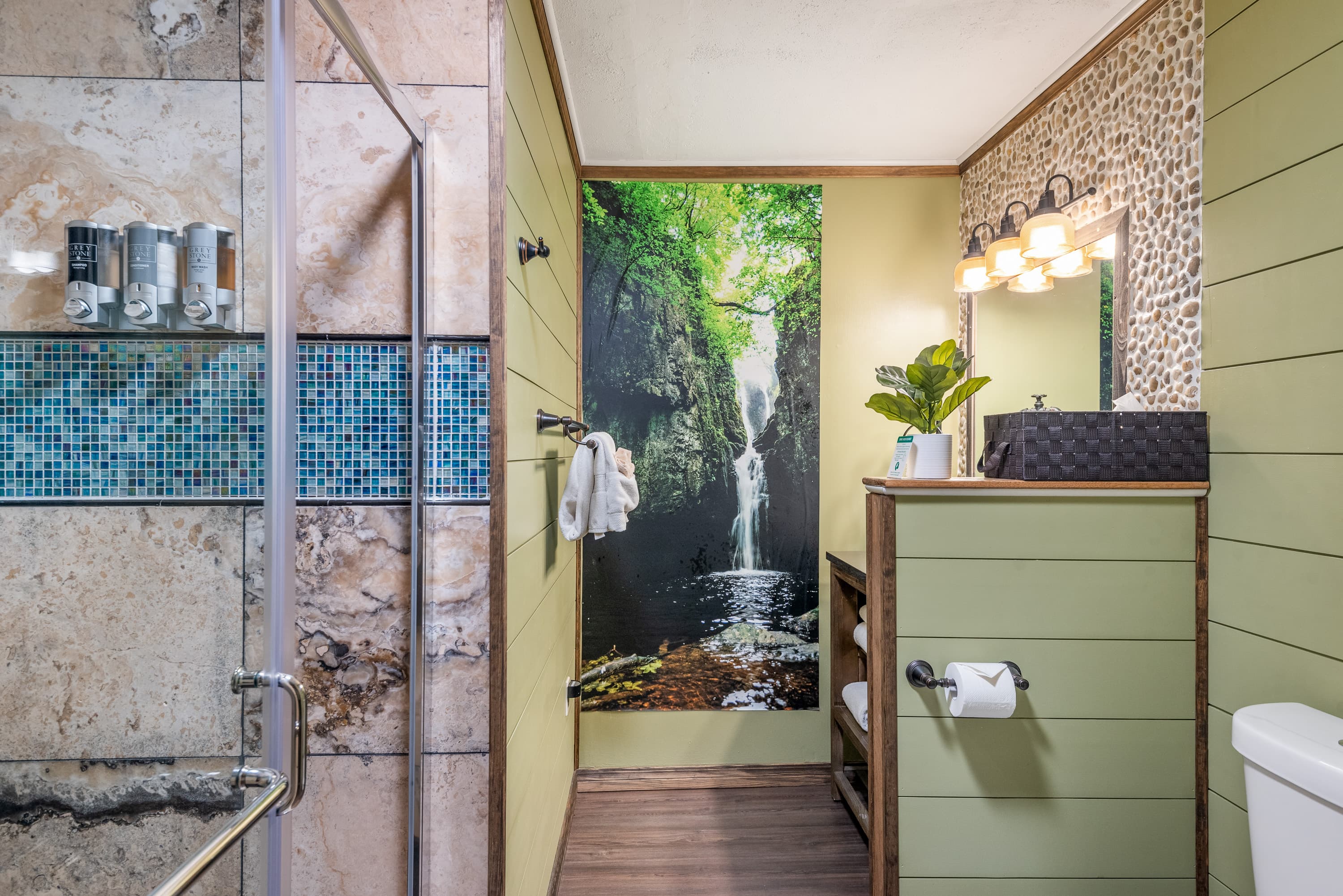 A rustic bathroom features a large wall mural of a waterfall, a walk-in shower with tiled walls, and a sink with a stone backsplash.