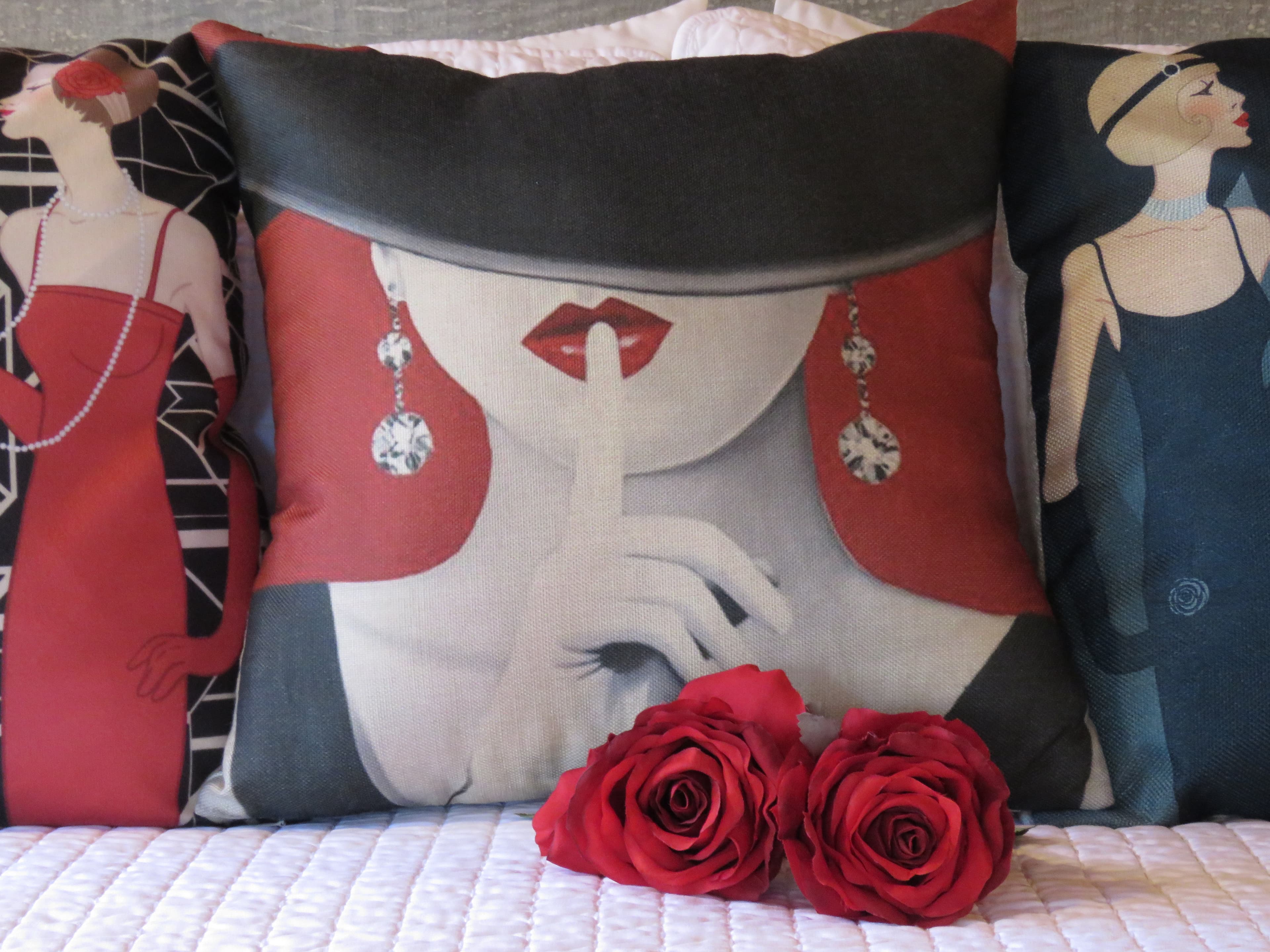 A close-up of a bed with decorative pillows and two red roses. The central pillow has a modern, artistic design of a woman with red hair, wearing a wide-brimmed hat, and holding a finger to her lips.
