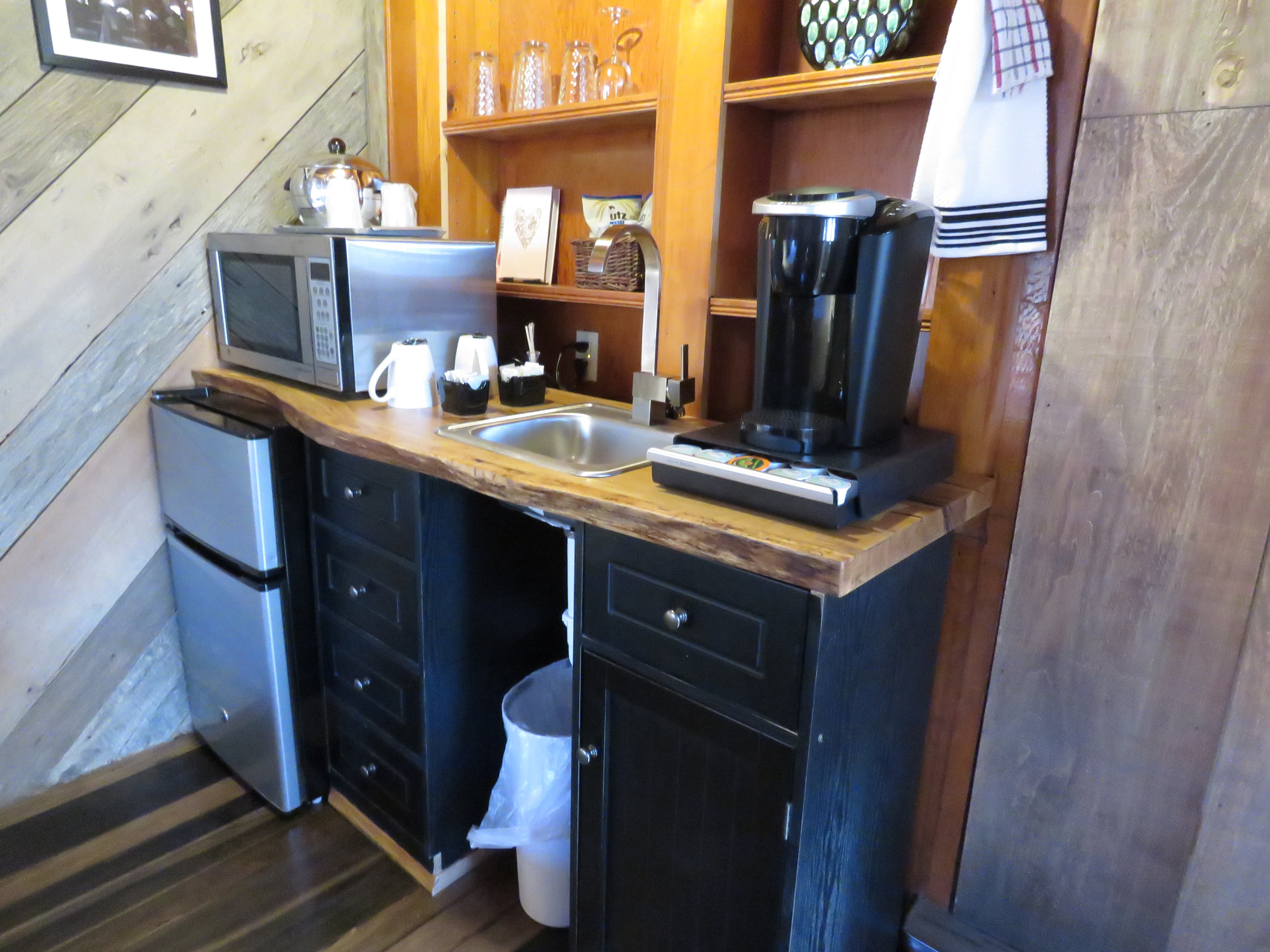 A small, rustic kitchenette with a live-edge countertop and black cabinets. The space has a microwave, a mini-fridge, and a coffee maker.