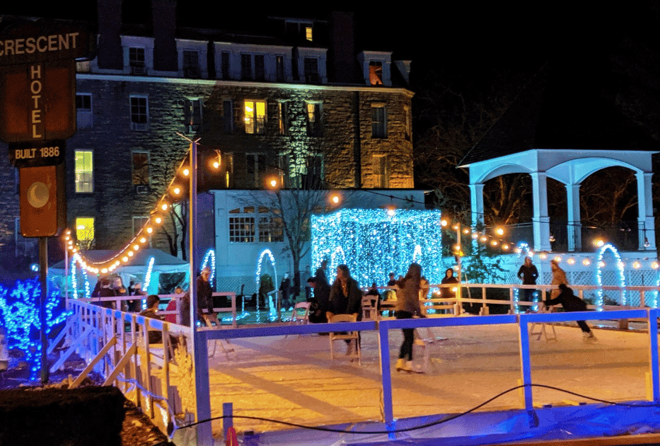 A festive outdoor ice skating rink is illuminated by colorful lights beside a historic hotel at night.