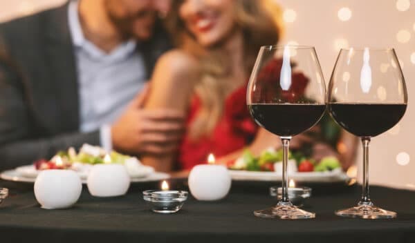 A romantic dinner setting with two glasses of red wine, candles, and a couple in the background.