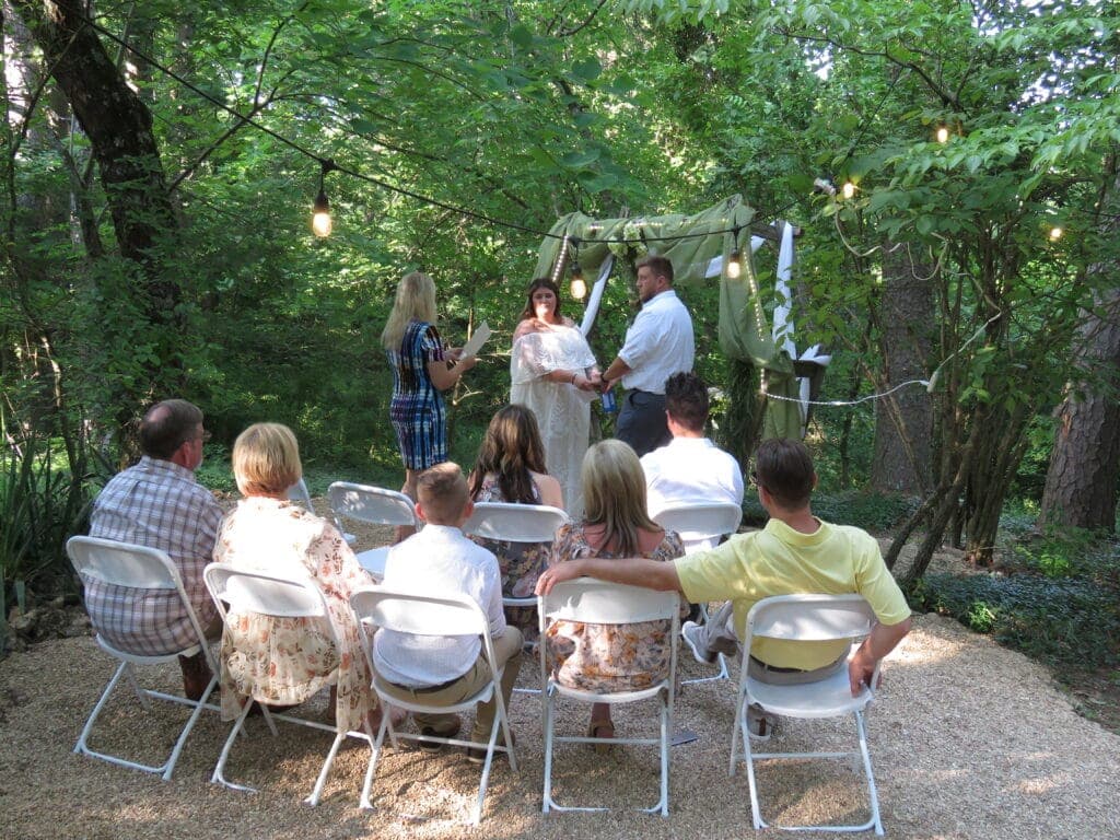 A couple exchanges vows under a floral arch in a wooded area, while guests sit in white chairs.