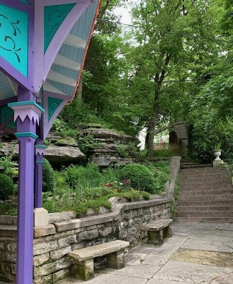 A colorful gazebo stands beside stone benches and lush greenery, leading to a staircase surrounded by rocky landscaping.