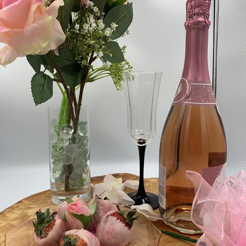 A bottle of rosé champagne, a flute glass, a vase of flowers, and decorated strawberries on a wooden tray.