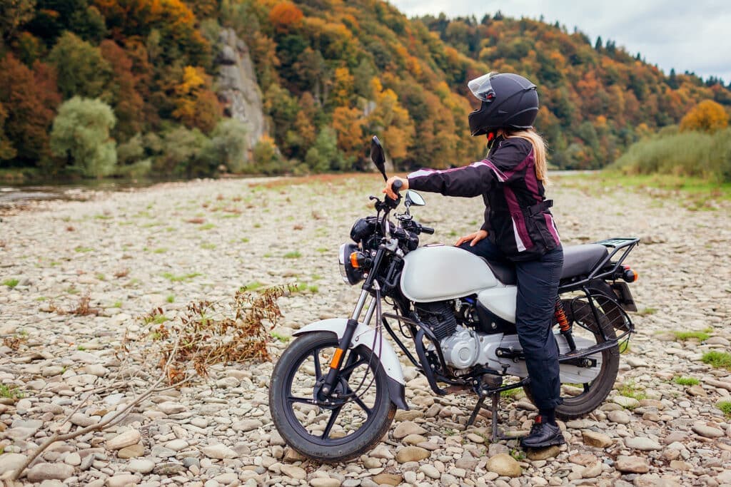A person in a motorcycle helmet stands next to a parked motorcycle on a rocky riverbank surrounded by autumn foliage.