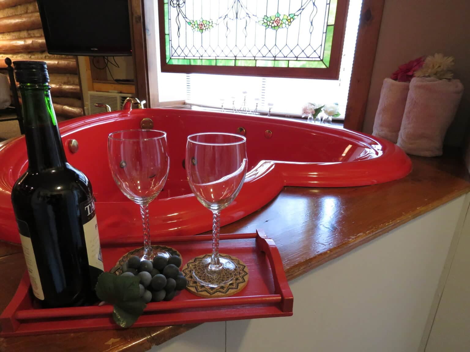 A red heart-shaped jacuzzi is accompanied by a bottle of wine and two glasses on a wooden tray.